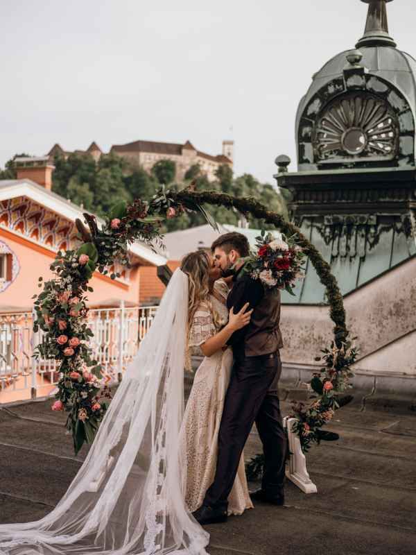 Bride and groom share a kiss under a circular floral arch on a rooftop with a castle backdrop.
