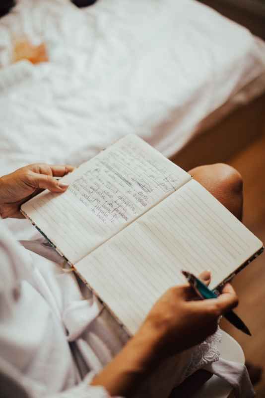 Bride in white robe writing wedding vows in a lined notebook with a pen before the ceremony.
