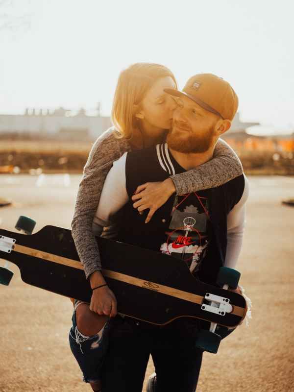A couple sharing a tender kiss outdoors, man holding a longboard skateboard, wearing casual clothes in warm sunset light.