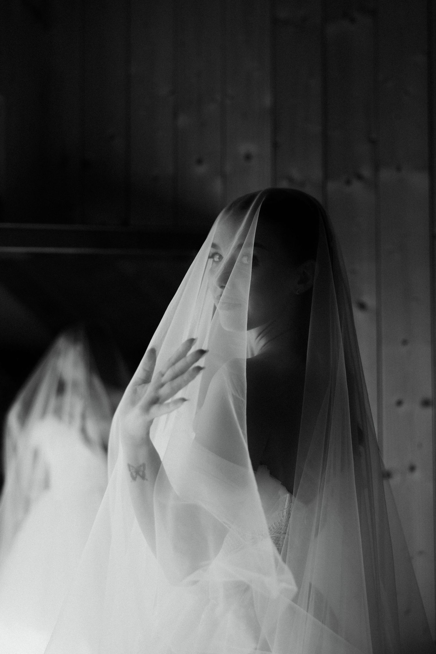 Black and white photo of a bride covered by a sheer veil, hand raised, gazing forward in a dark wooden interior.