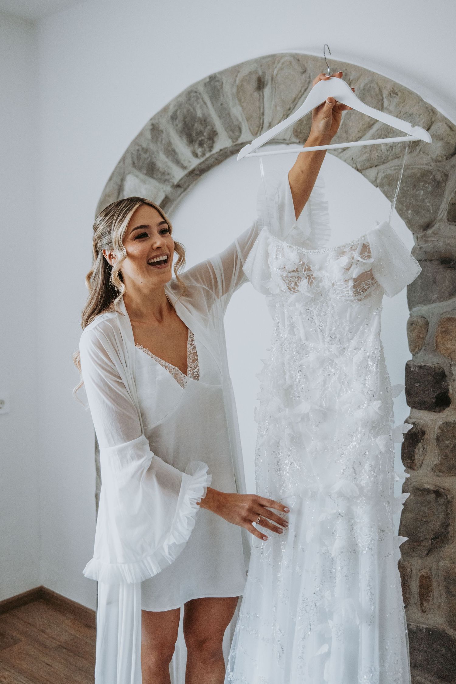 Smiling bride in white robe holding up her elegant lace wedding dress on a hanger in a bright room.