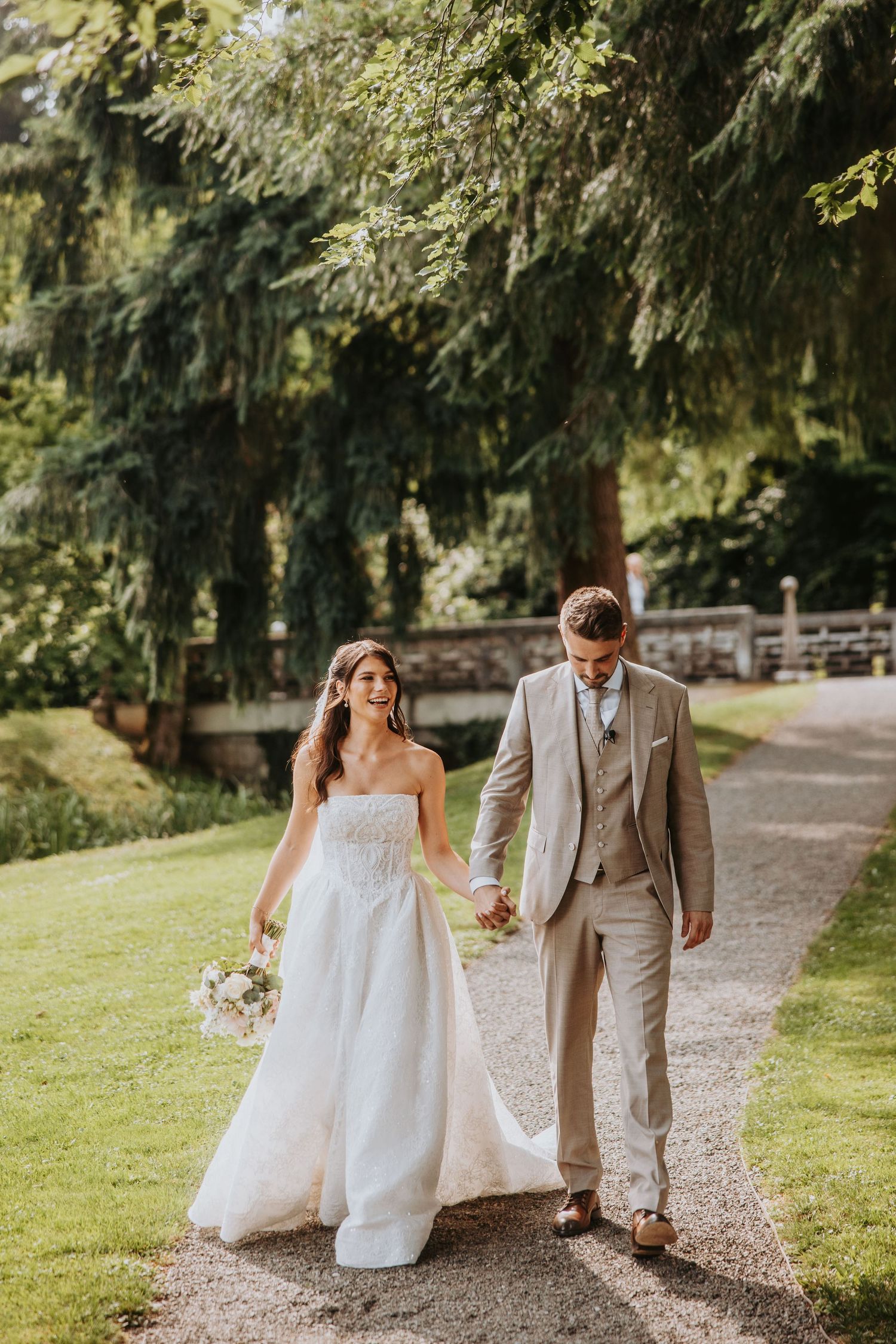 Bride in white gown and groom in tan suit walk hand-in-hand along sunlit garden path.