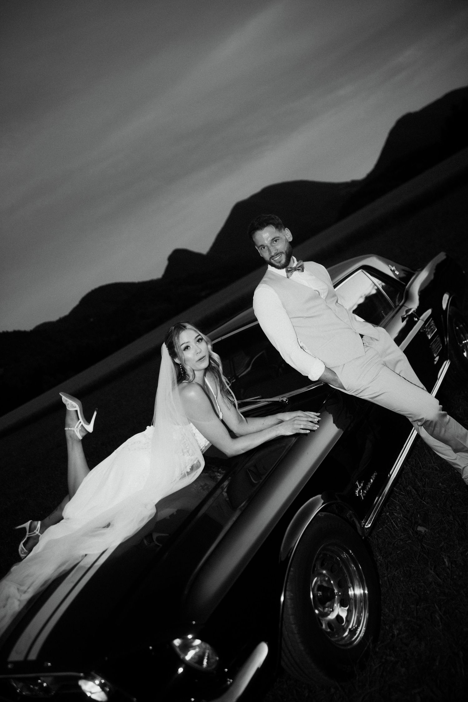 Black and white photo of bride and groom posing with a classic vintage car at dusk in mountains.