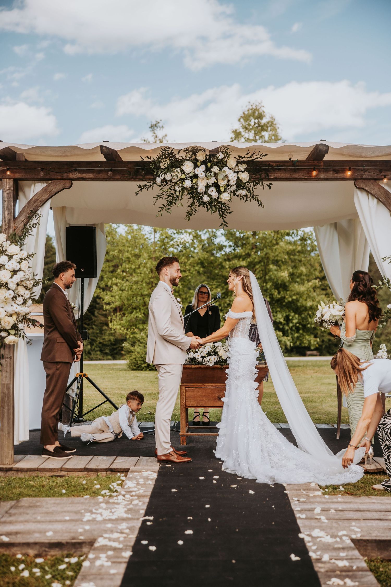 Bride and groom exchange vows under floral wooden arch with white roses during outdoor wedding ceremony.