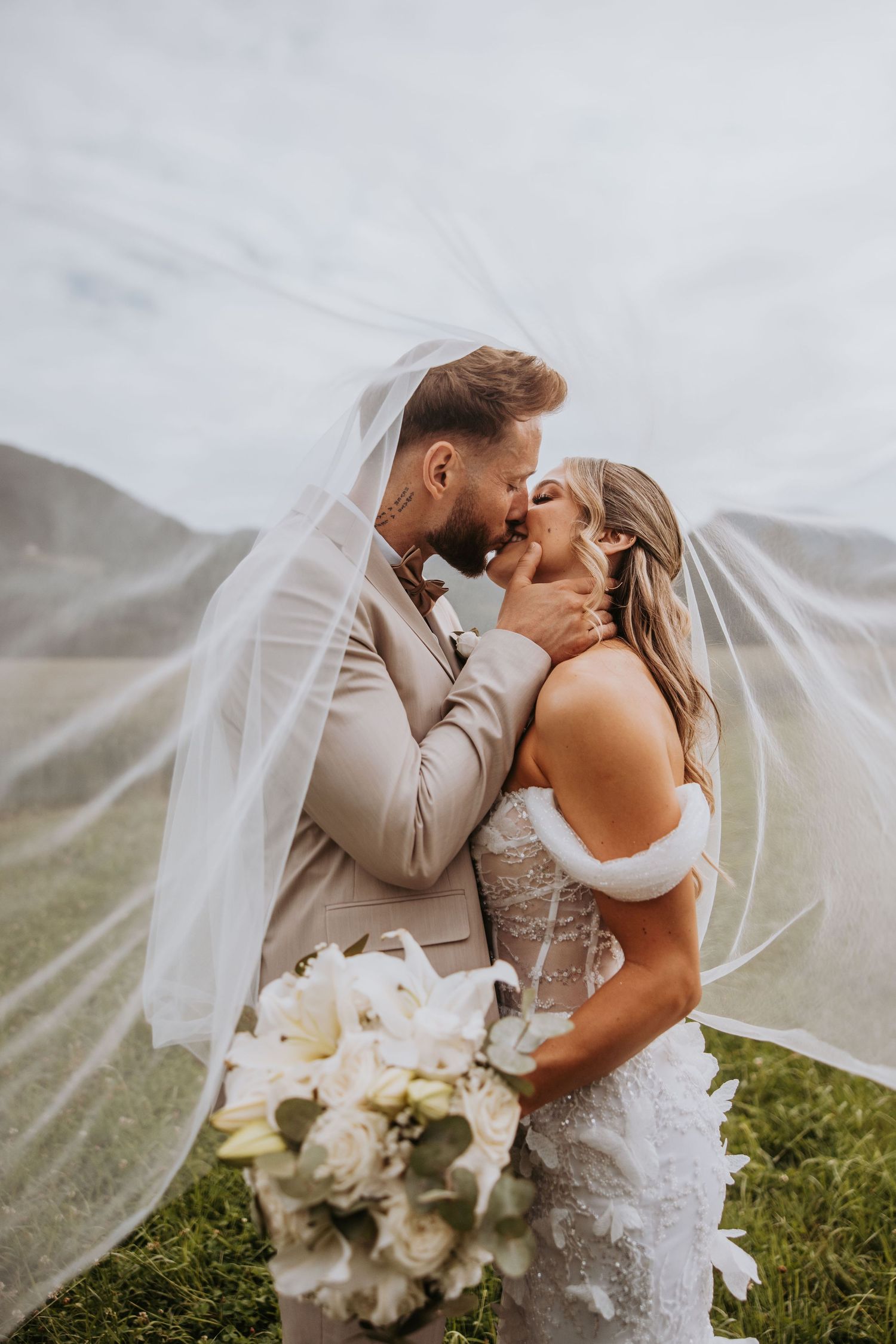 Bride and groom kissing under flowing veil with mountain backdrop and white lily bouquet.