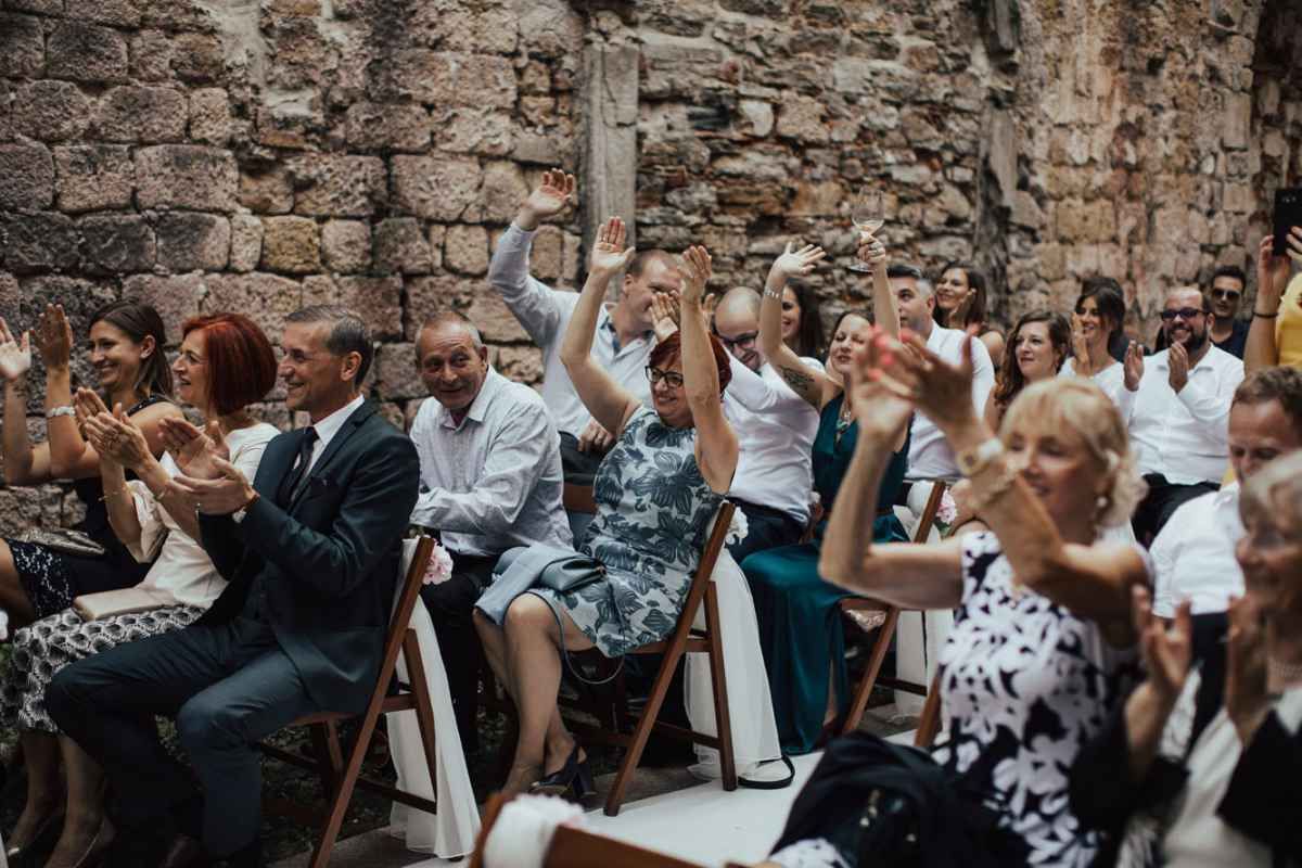 Wedding guests clapping and celebrating during an outdoor ceremony against a rustic stone wall backdrop.