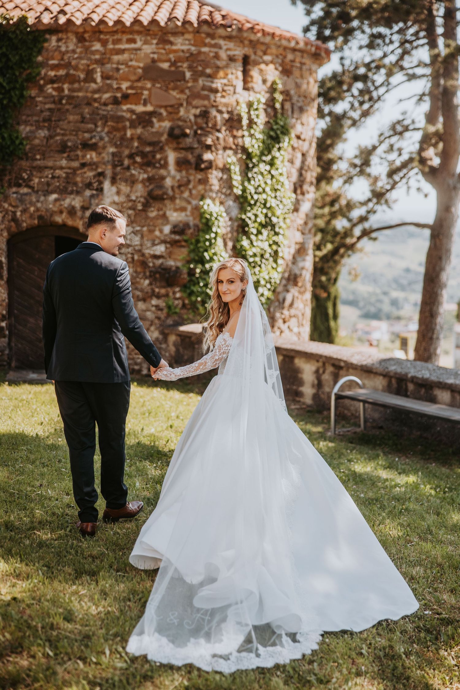 Bride and groom holding hands near rustic stone castle in lush garden on wedding day.