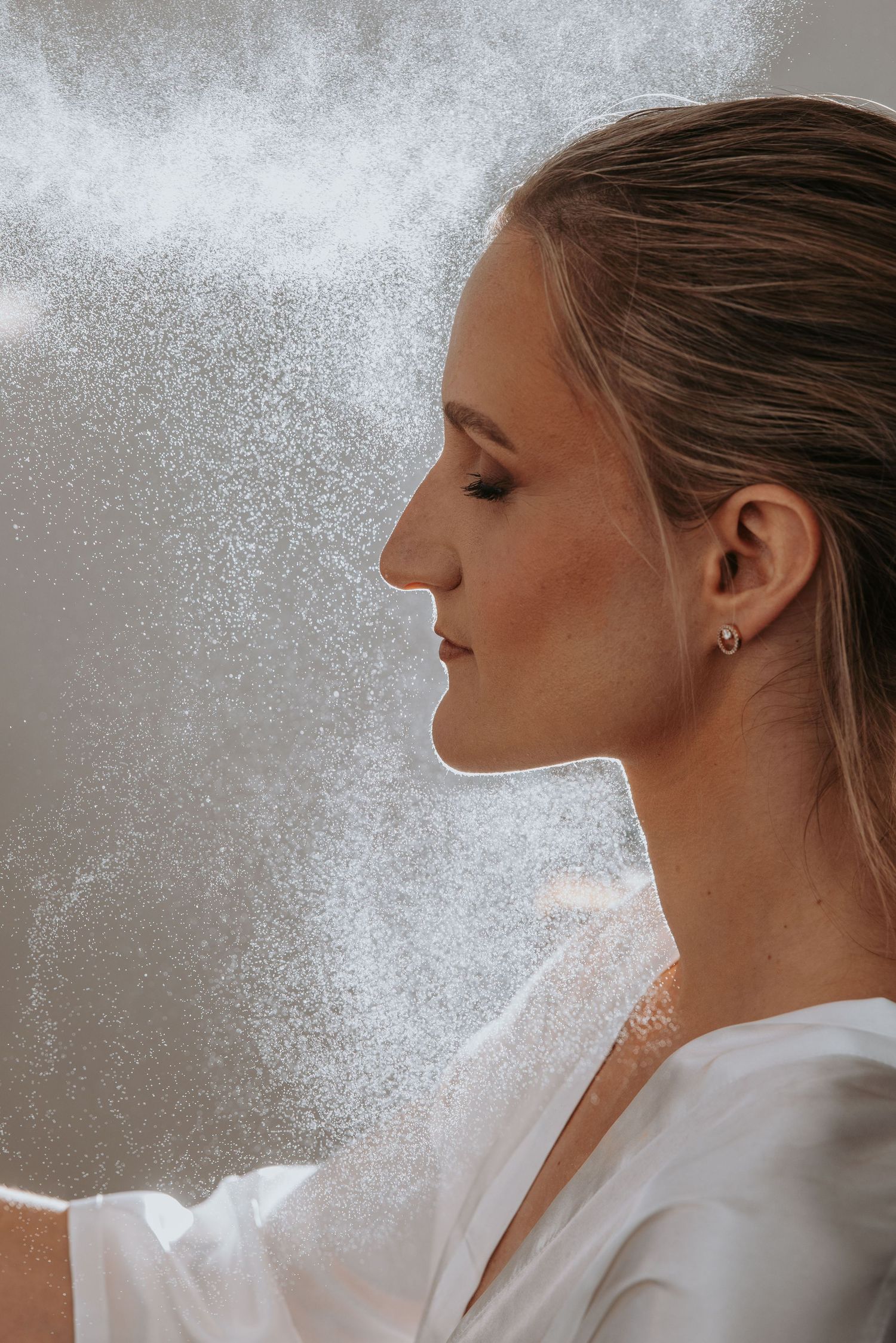 Young woman in profile view with glowing skin, wearing a white top and hoop earring, surrounded by mist spray.
