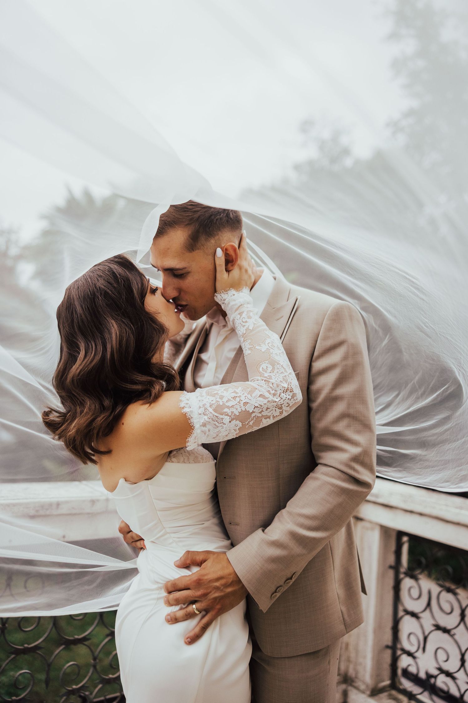 Bride and groom sharing romantic kiss under flowing white veil on balcony, groom in beige suit.