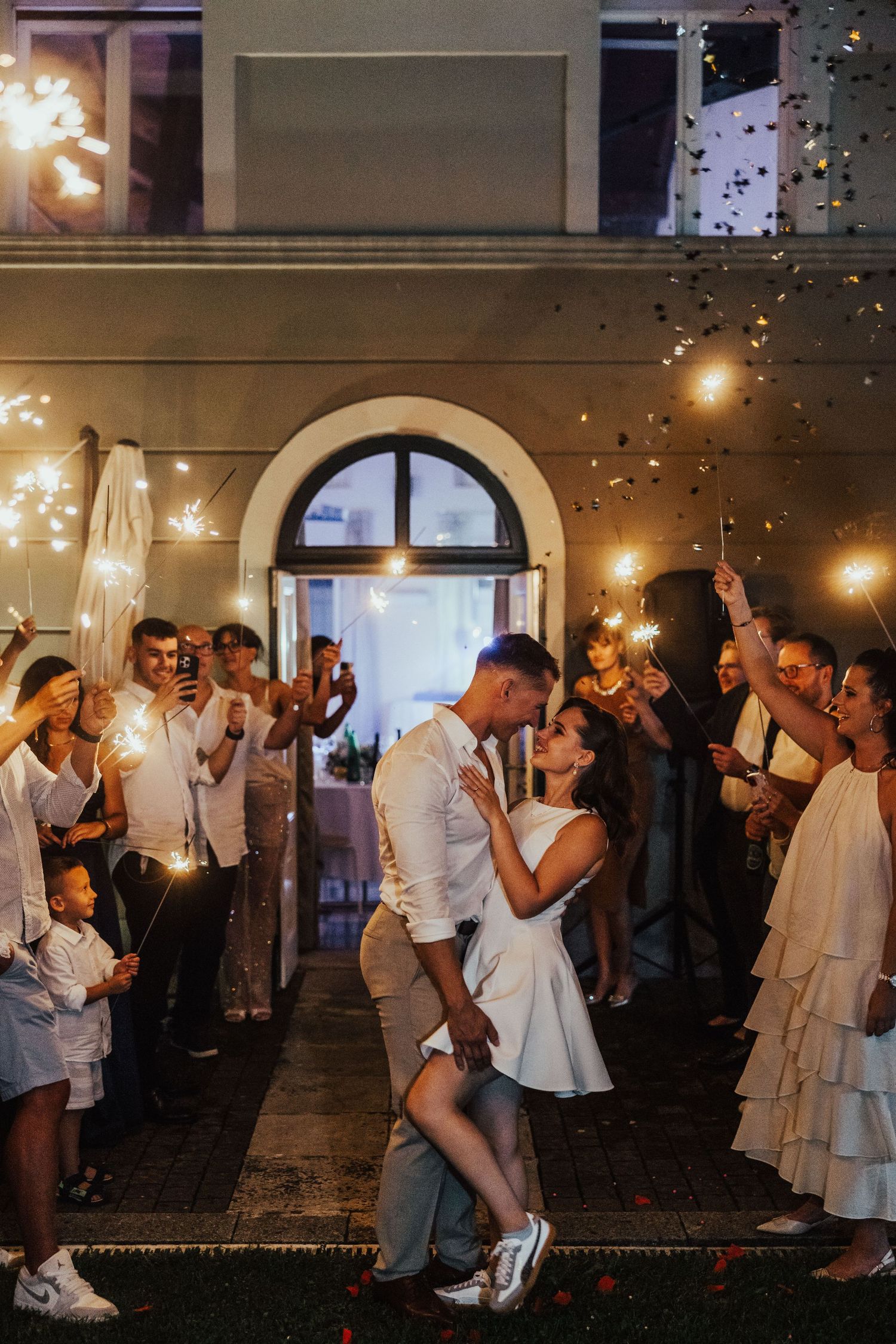 Couple sharing a romantic kiss surrounded by guests holding sparklers and confetti at an outdoor wedding celebration at night.