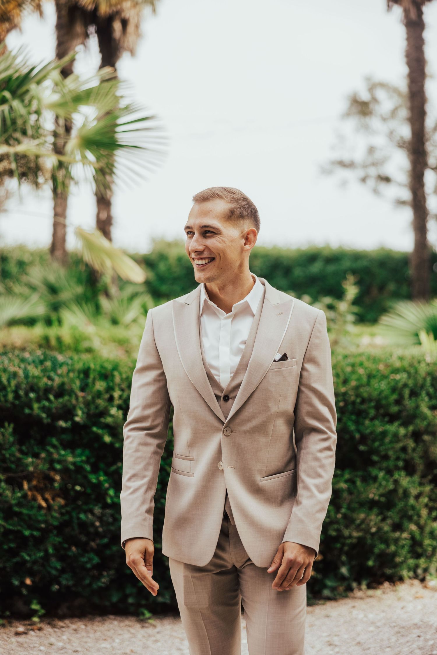 Smiling groom in a beige suit stands outdoors among palm trees and lush greenery on his wedding day.