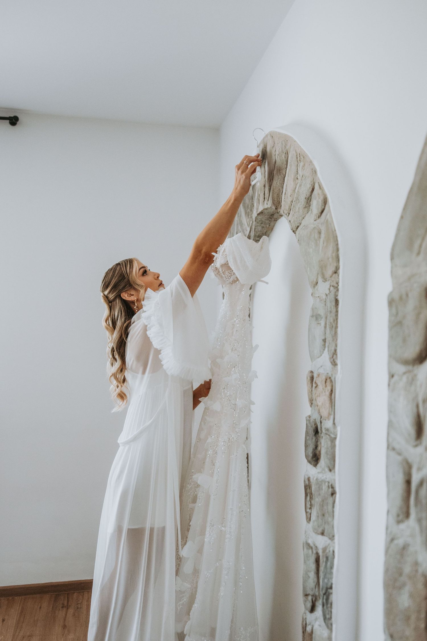 Bride in white robe reaching up to adjust her wedding dress hanging on a stone arch mirror.