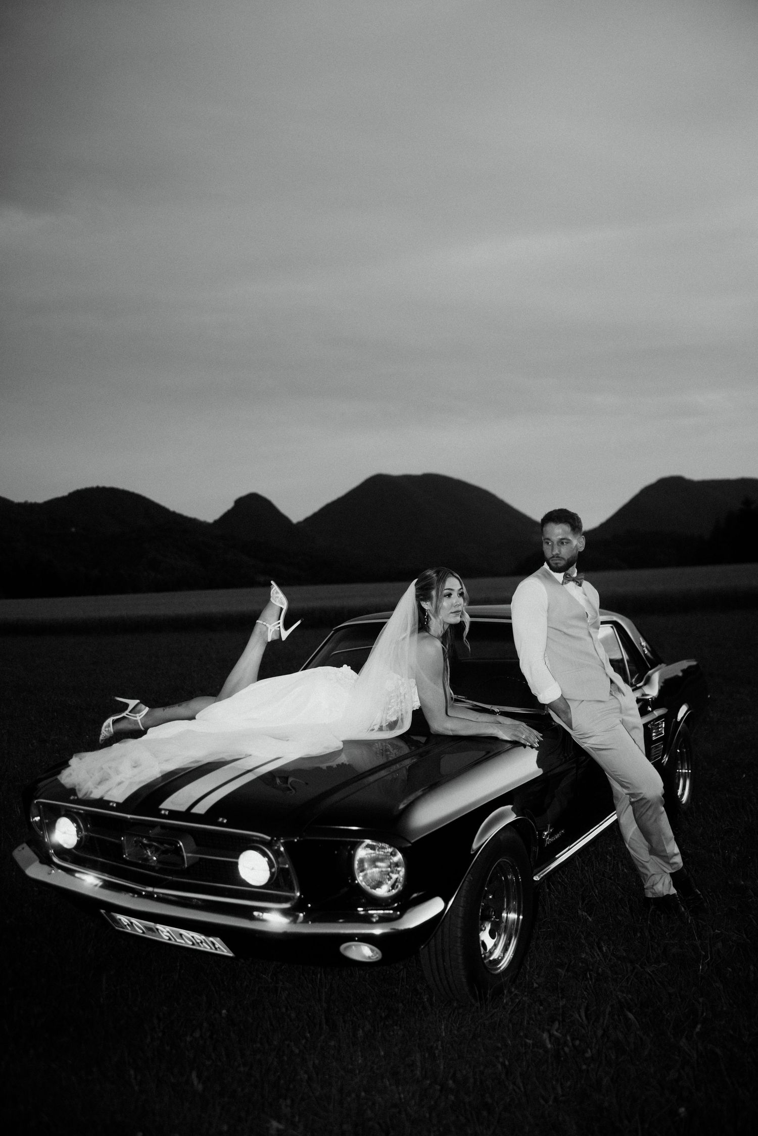 Black and white photo of bride and groom leaning on a classic Mustang with mountain backdrop at dusk.