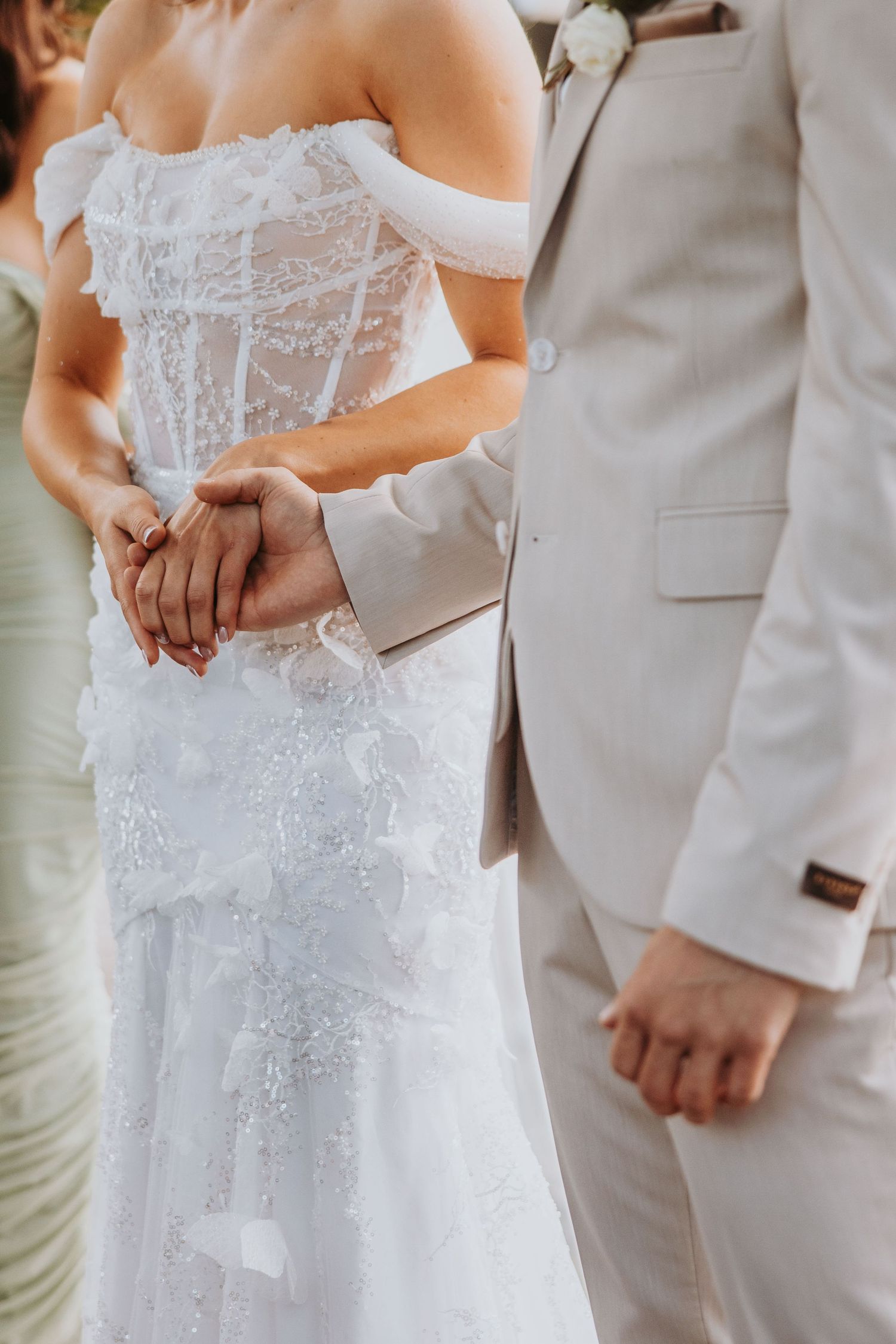 Close-up of bride and groom holding hands, bride in lace corset gown, groom in beige suit.