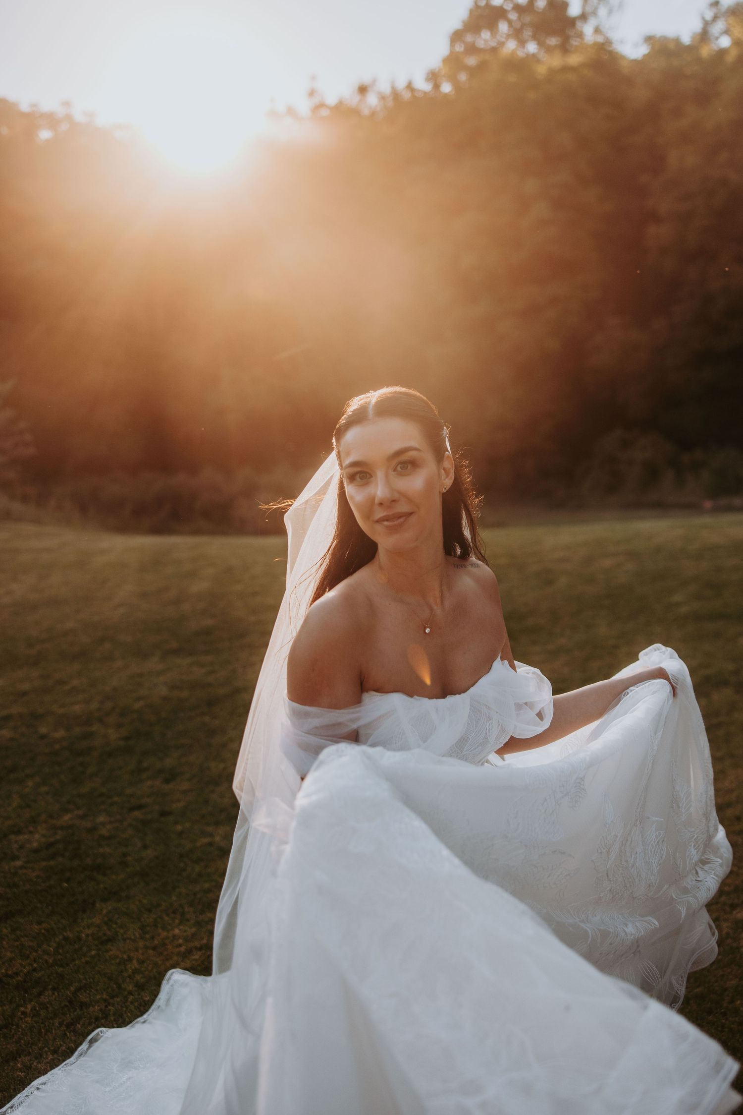 Radiant bride in an off-shoulder white wedding dress twirling on a green lawn at sunset.