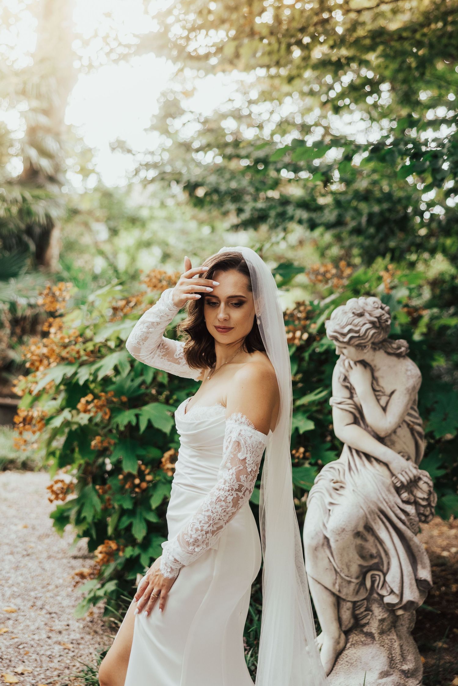 Bride in lace wedding dress and veil poses beside stone statue in lush garden with soft sunlight.