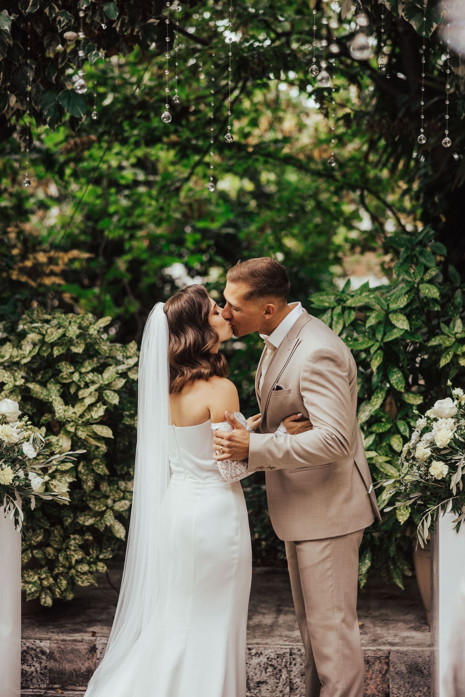 Bride and groom share first kiss under floral arch surrounded by lush greenery at outdoor ceremony.