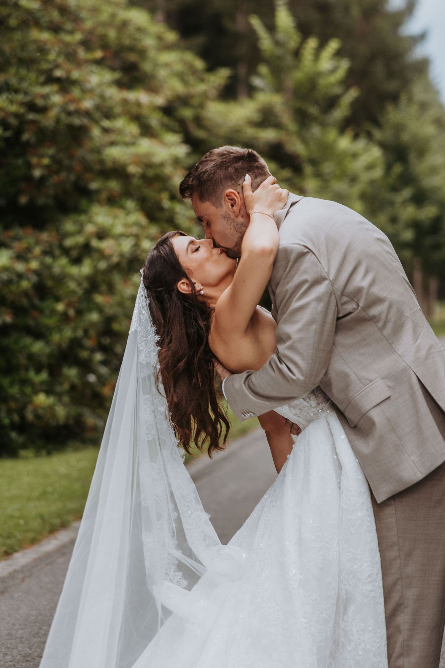 Groom dips bride for romantic kiss on tree-lined path in lush garden setting.