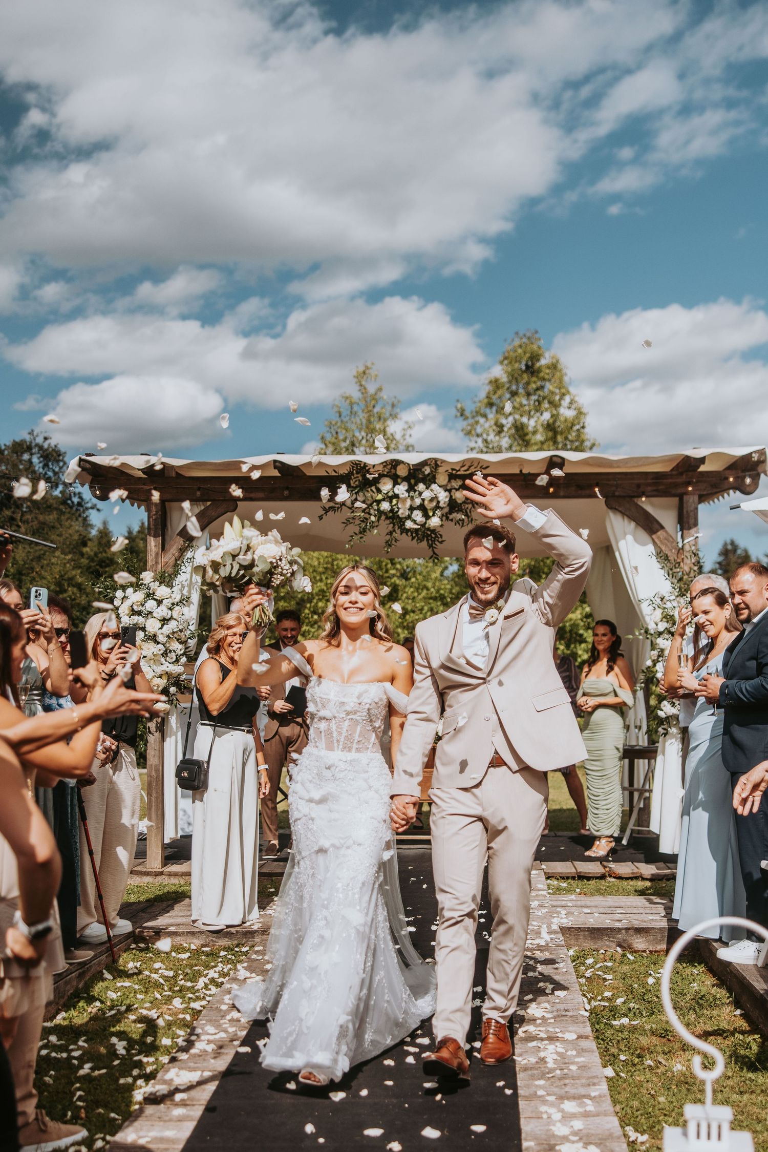 Newlyweds celebrate recessional as guests toss white petals under blue sky at outdoor ceremony.