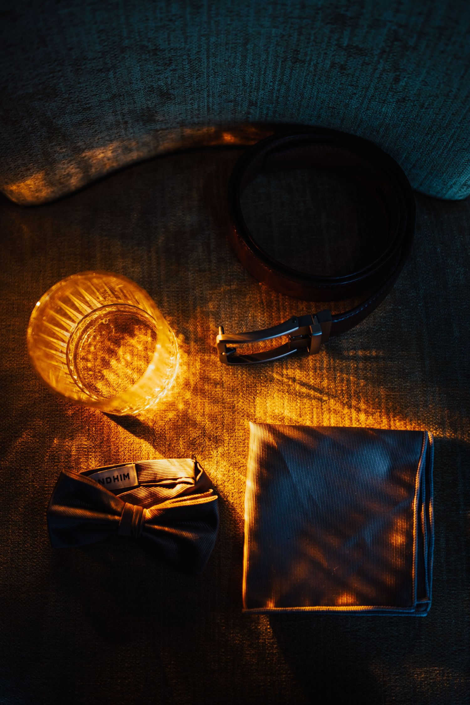 Grooms wedding accessories including belt, bow tie, pocket square, and whiskey glass in dramatic golden light.