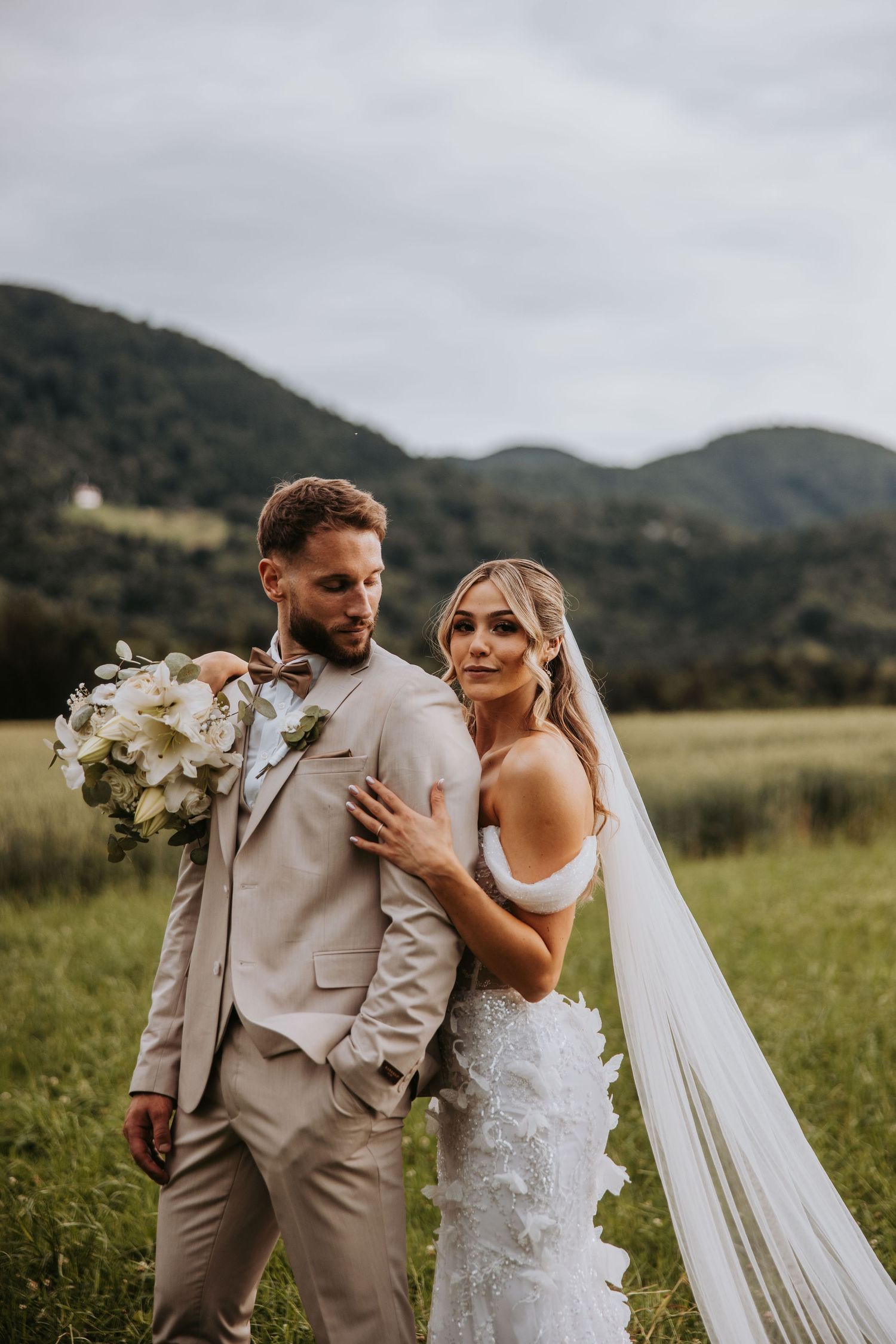 Bride and groom posing in green mountain meadow, groom in beige suit holding bouquet, bride in lace gown with veil.