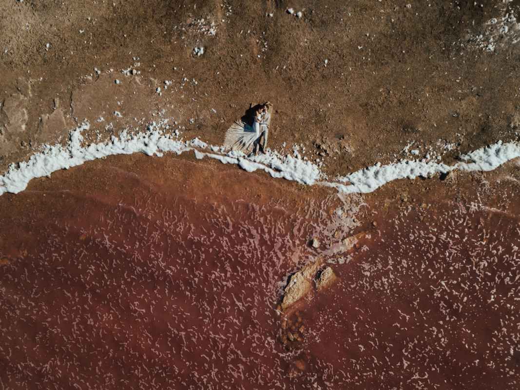 Aerial drone view of a couple standing at the edge of a red sandy beach with white ocean waves.