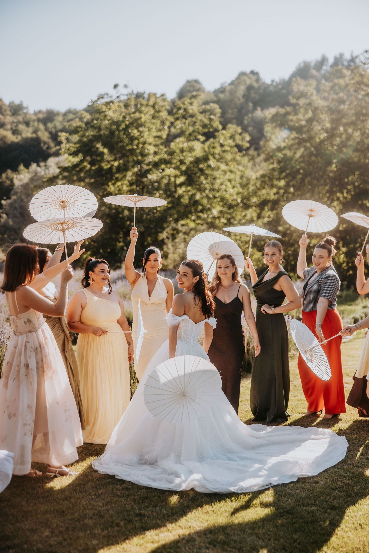Bride and bridesmaids in colorful gowns holding white parasols outdoors on sunny day.