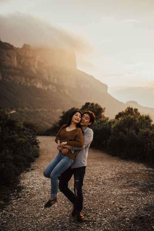 Couple embracing at sunset on a mountain trail, man kissing woman's cheek as golden light illuminates rocky peaks behind them.