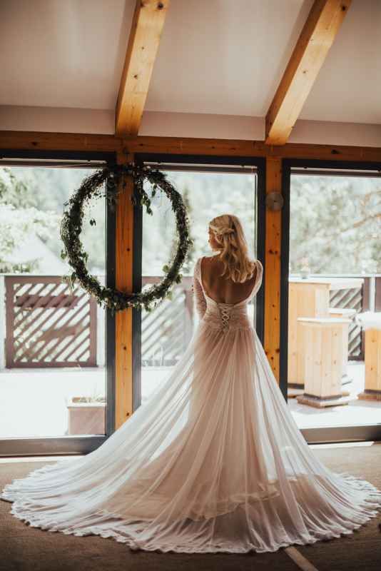 Bride in elegant backless lace gown standing before circular greenery wreath in rustic venue.