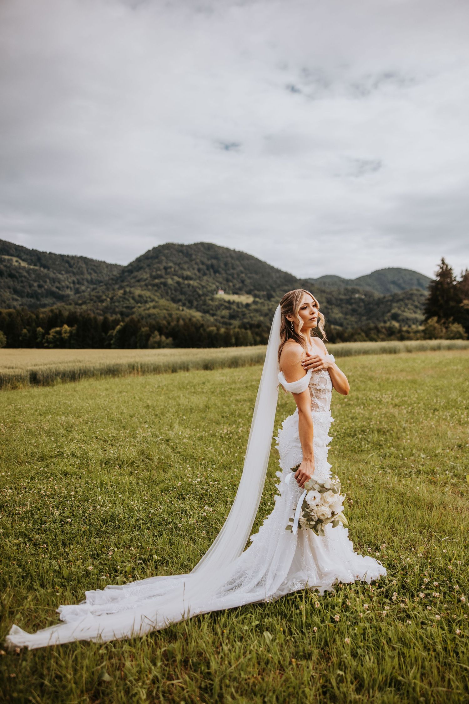 Full-length bridal portrait in lace mermaid gown with long flowing veil in a scenic mountain field.