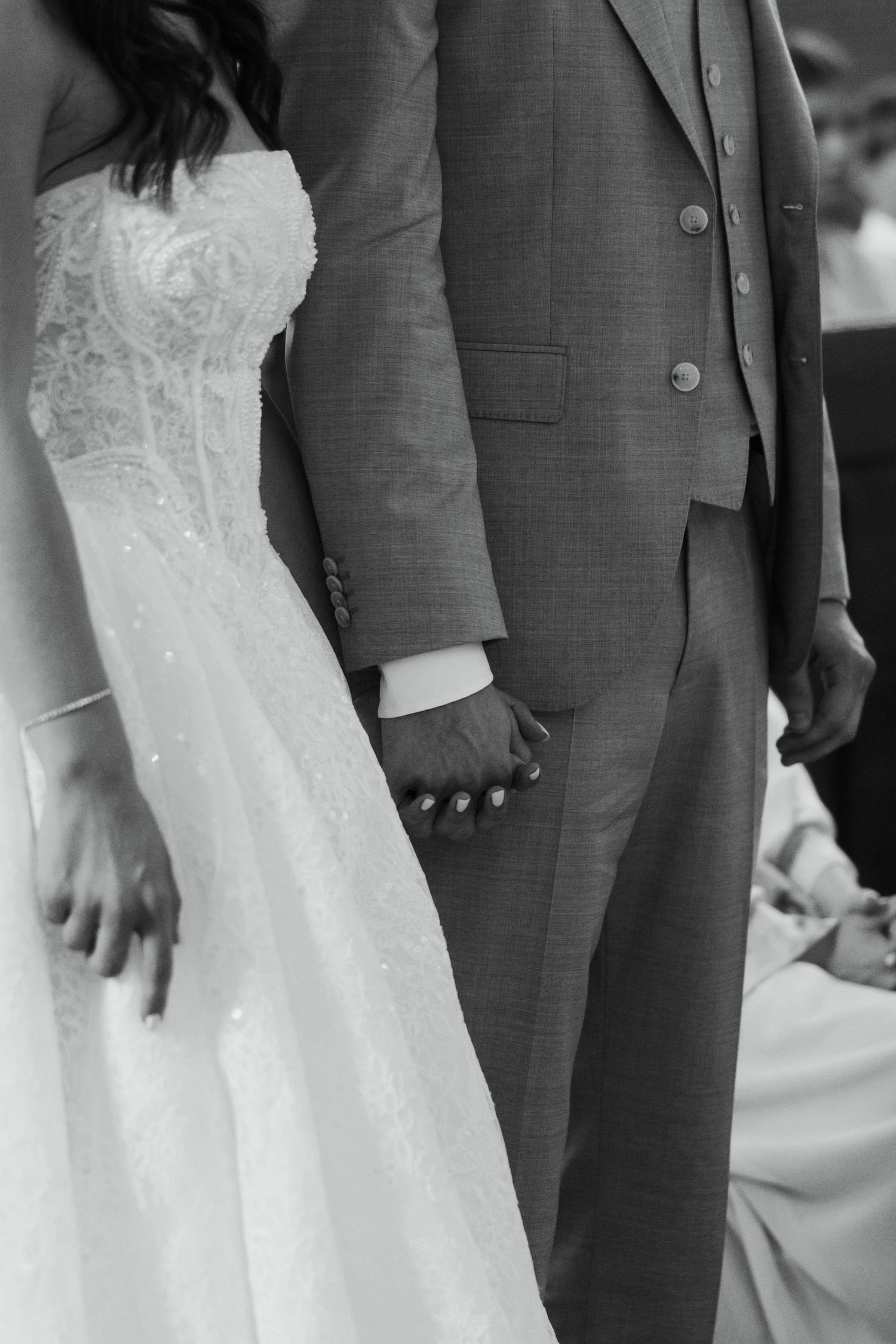 Black and white photo of bride and groom holding hands, bride in lace gown, groom in gray suit.