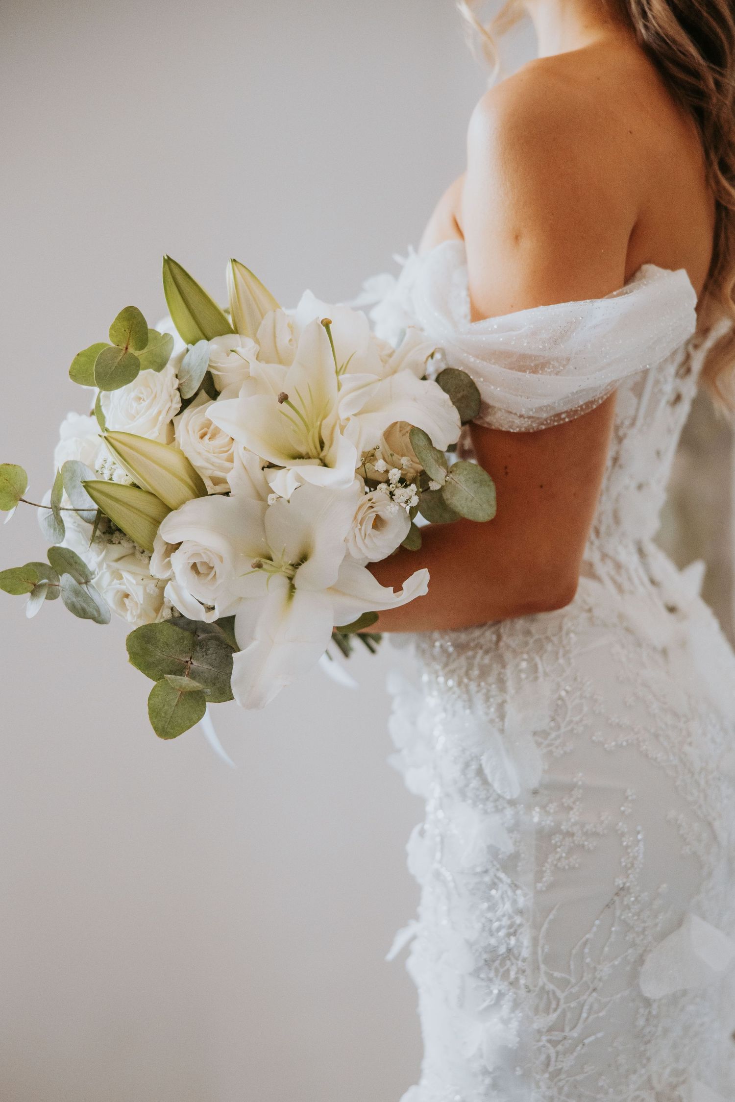 Bride in lace off-shoulder gown holding elegant white lily and rose bouquet.