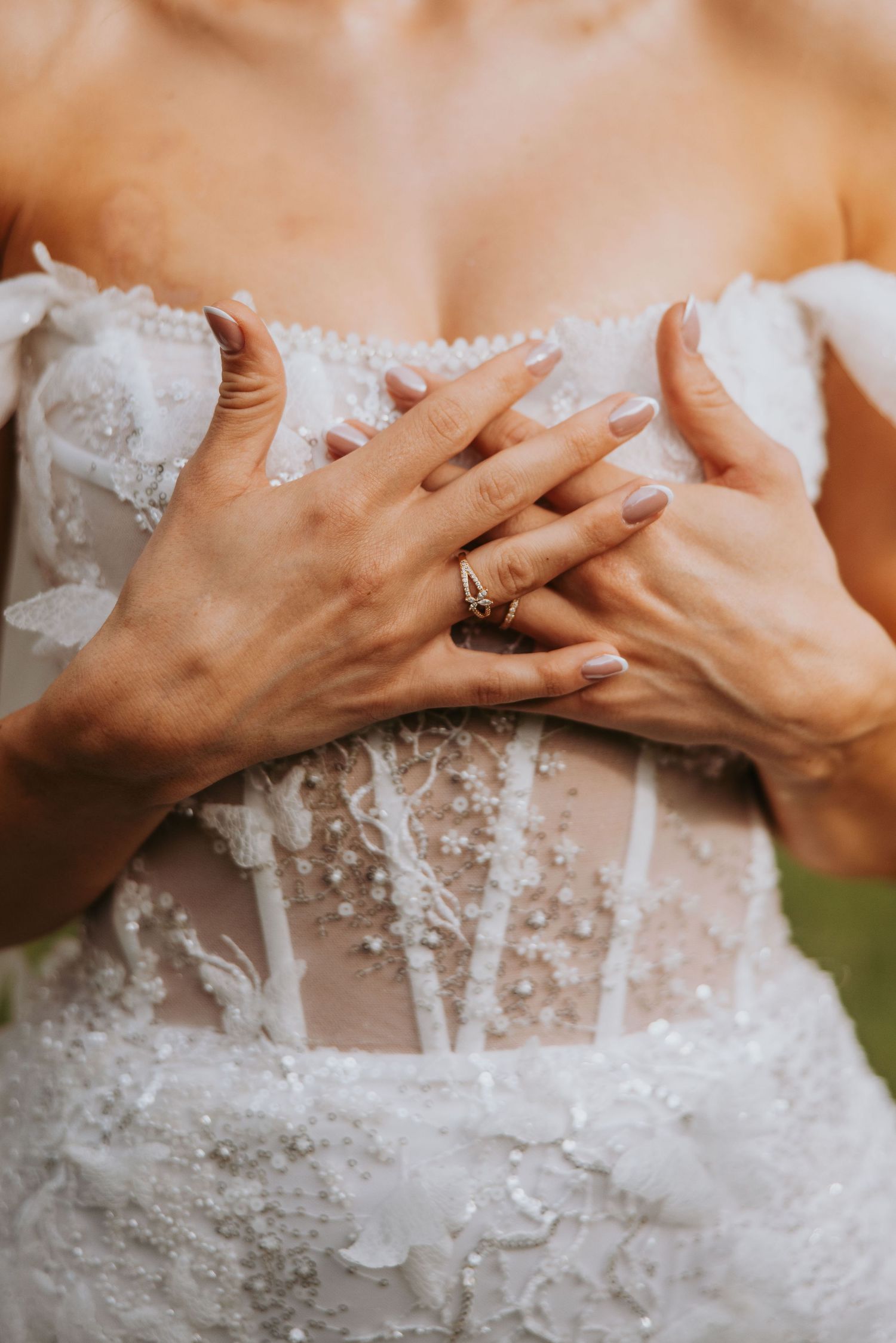Close-up of bride\u2019s hands with engagement ring resting on beaded lace corset wedding dress.
