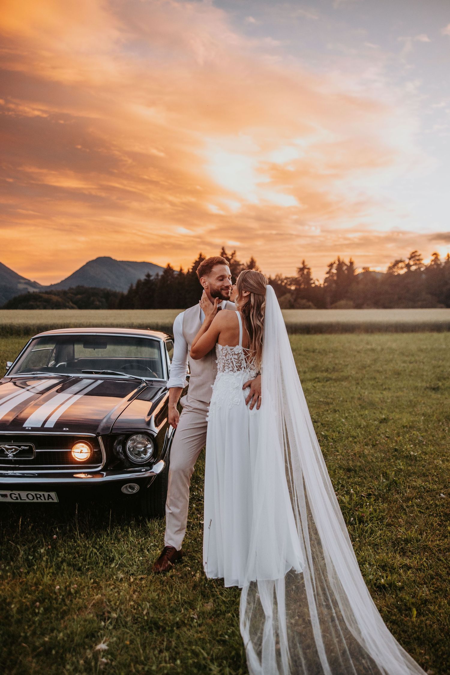 Bride and groom share romantic moment beside vintage Mustang at golden sunset in mountain meadow.