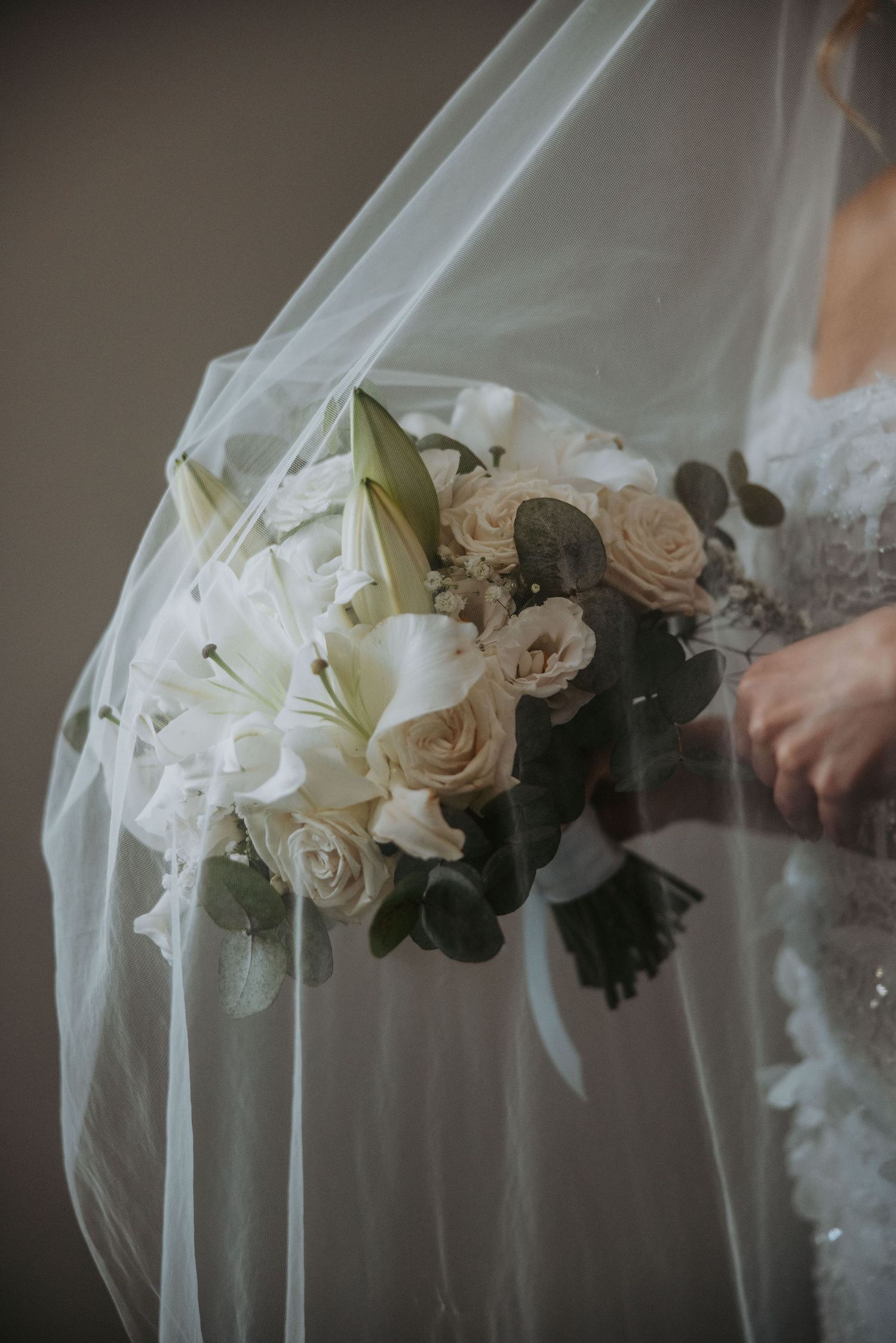 Bride holding white floral bouquet with lilies and roses beneath flowing wedding veil.