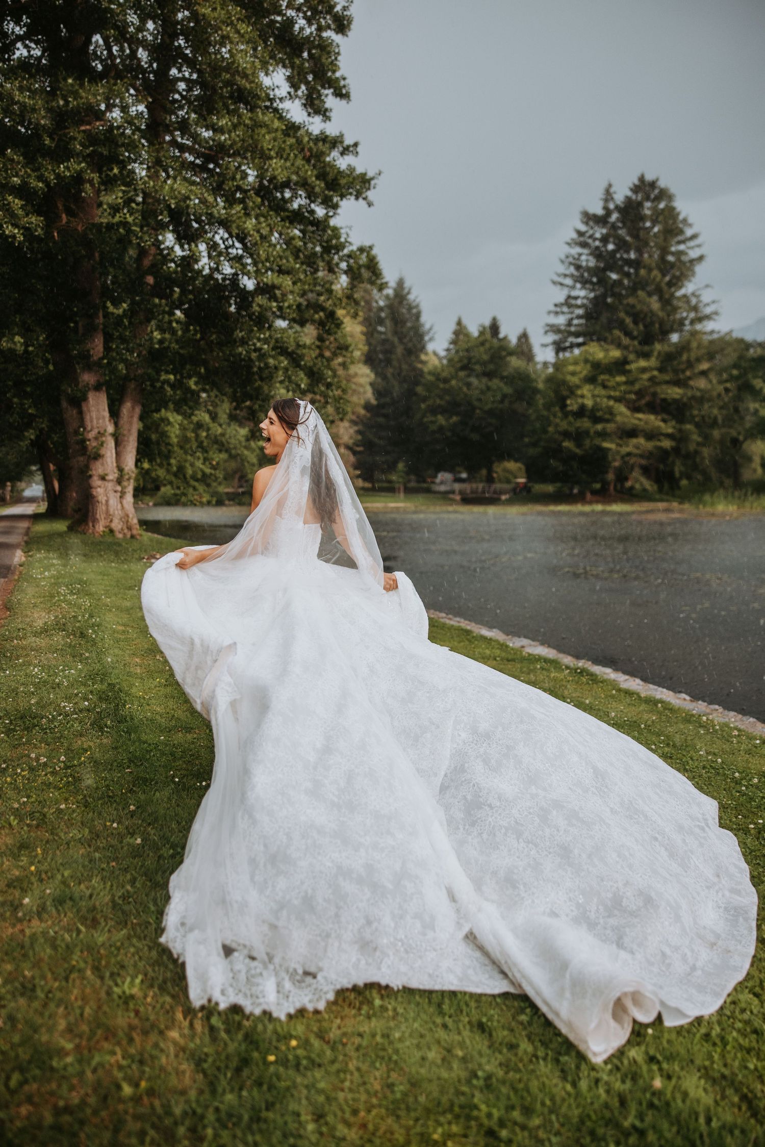 Bride in flowing white lace gown and veil poses gracefully on green lawn with trees and pond backdrop.