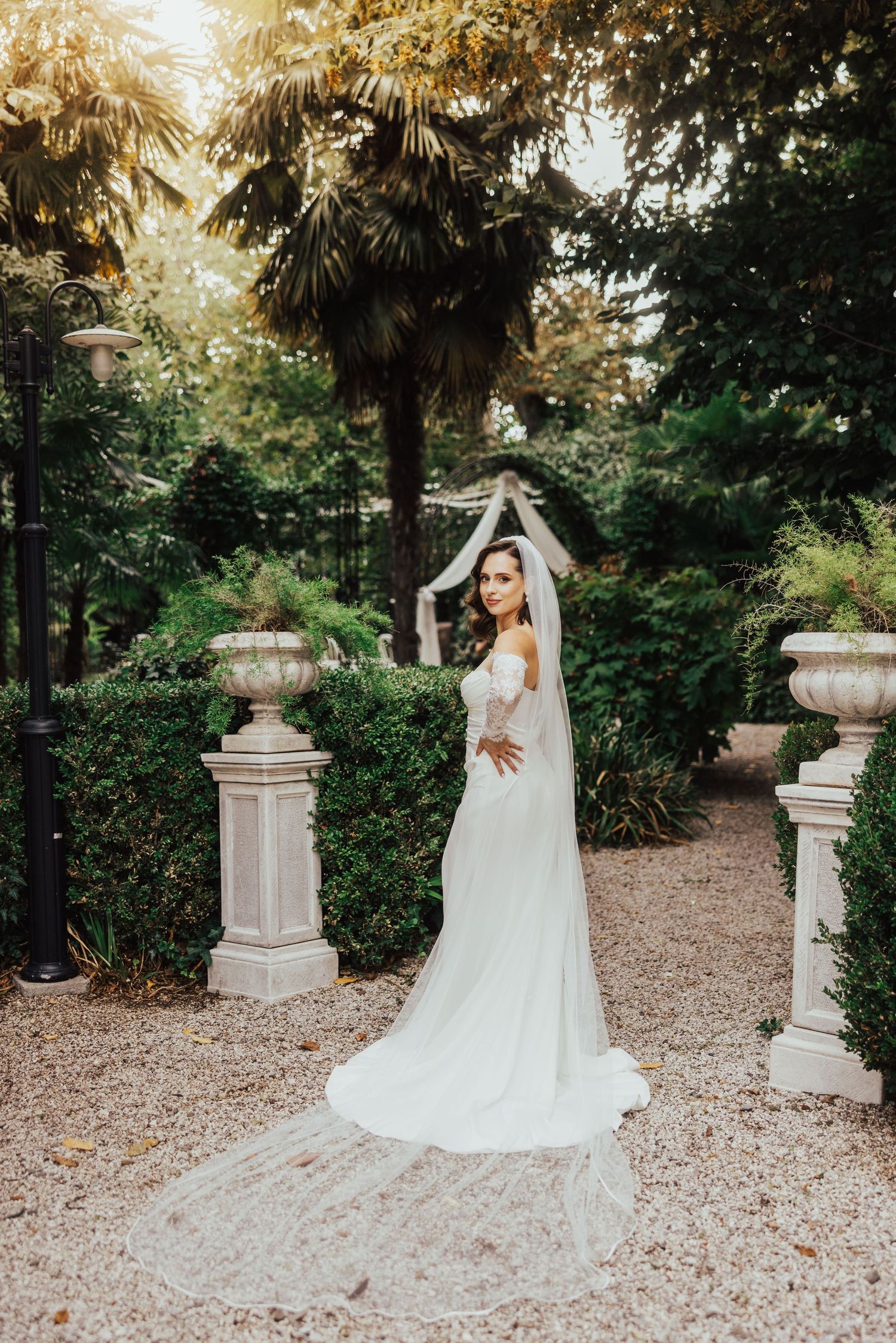 Bride in fitted white lace gown with cathedral veil posing in sunlit garden with stone columns.