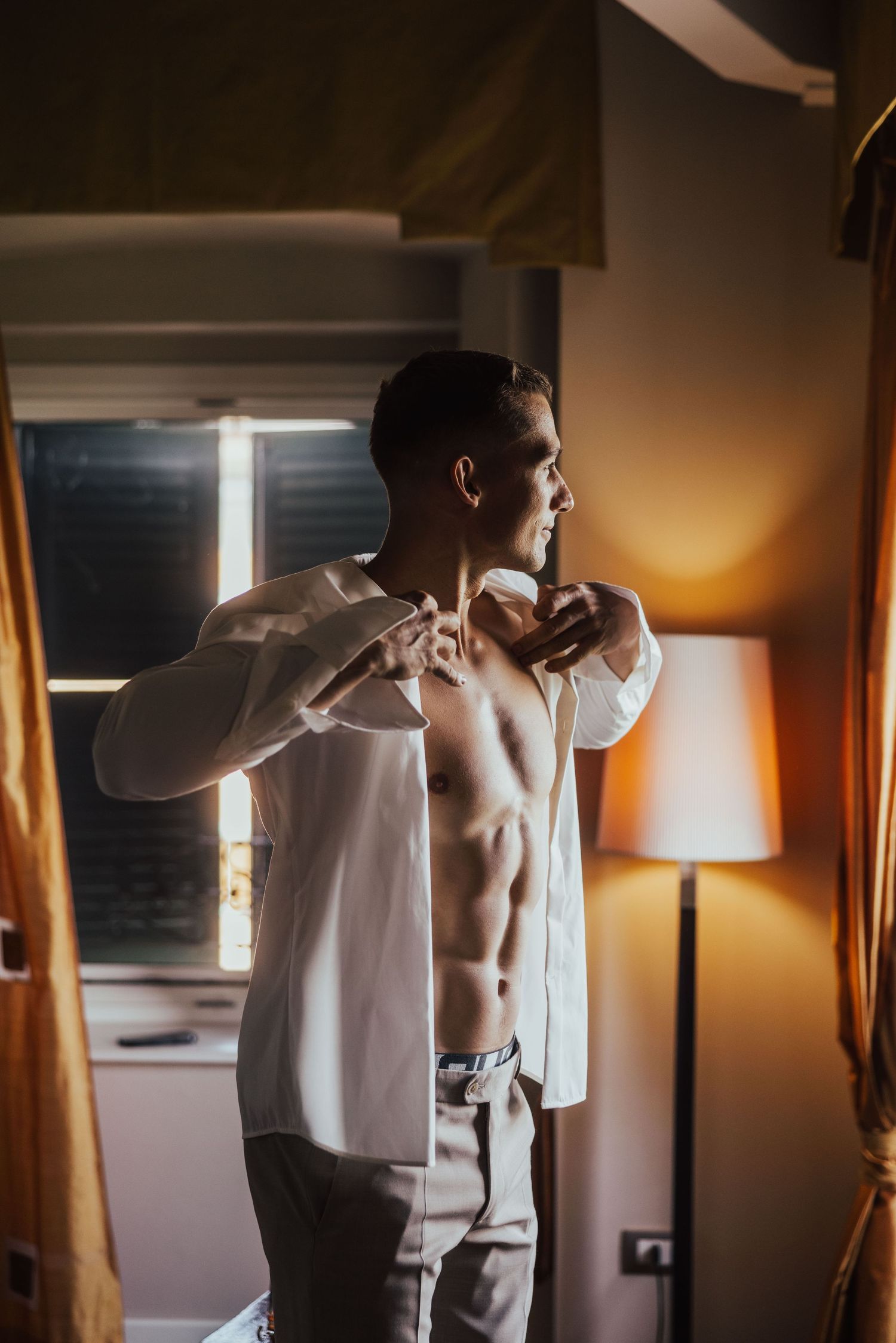 Fit groom buttoning white dress shirt in warmly lit hotel room while getting ready for wedding ceremony.
