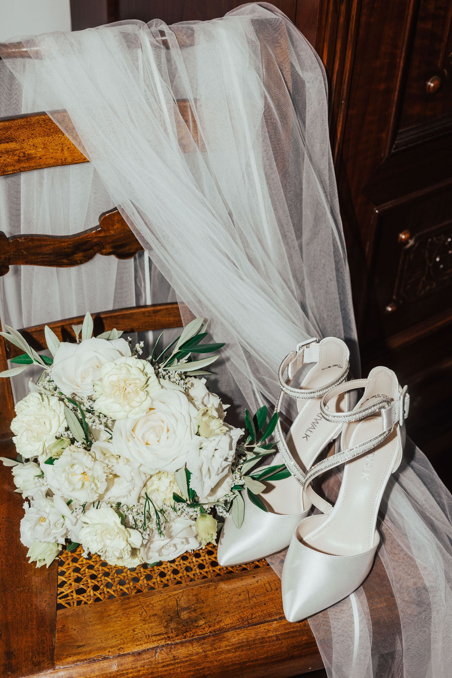 Bridal accessories with white rose bouquet, heels, and veil on wooden chair.