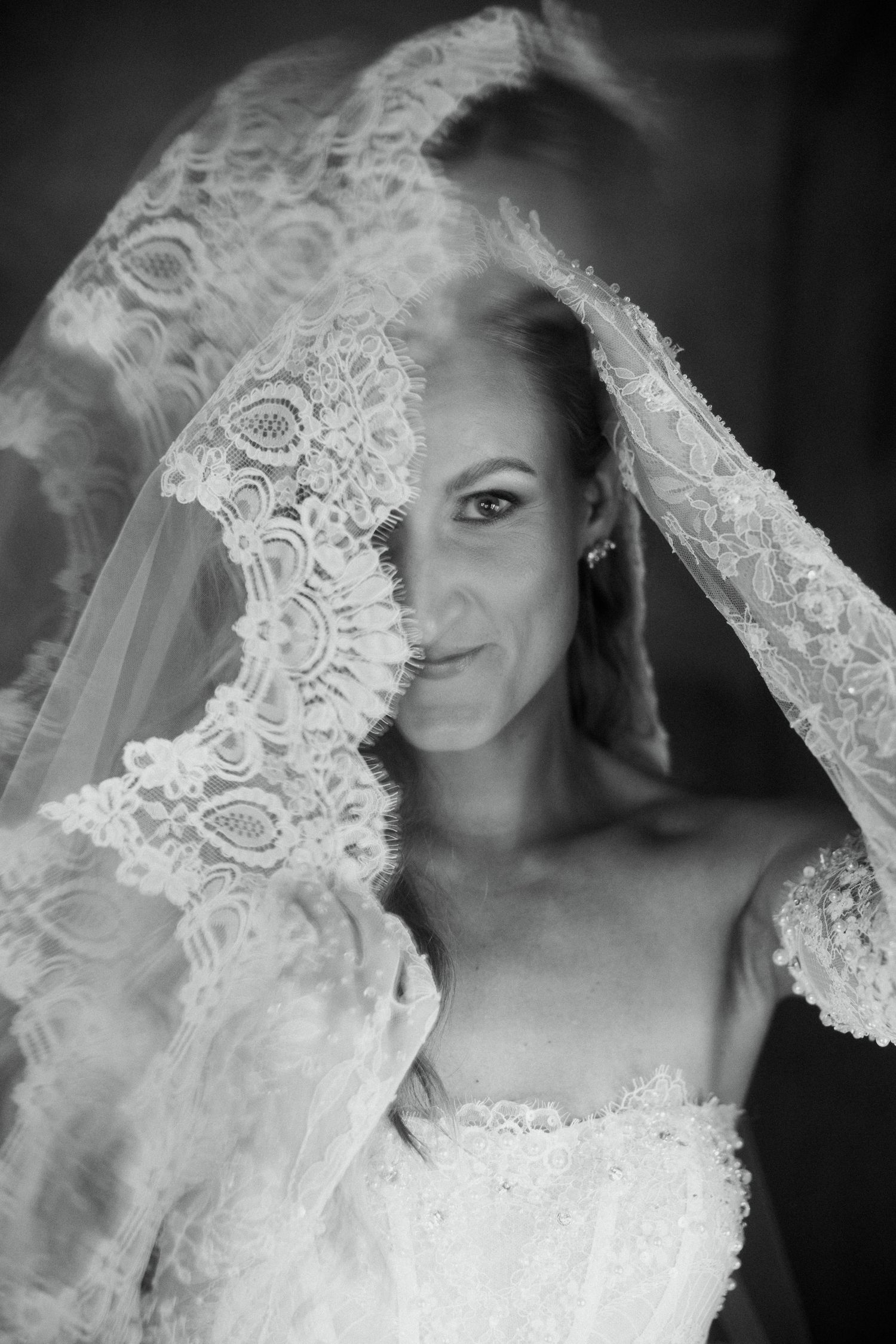 Black and white portrait of a bride lifting her lace veil, revealing her elegant wedding dress and serene smile.