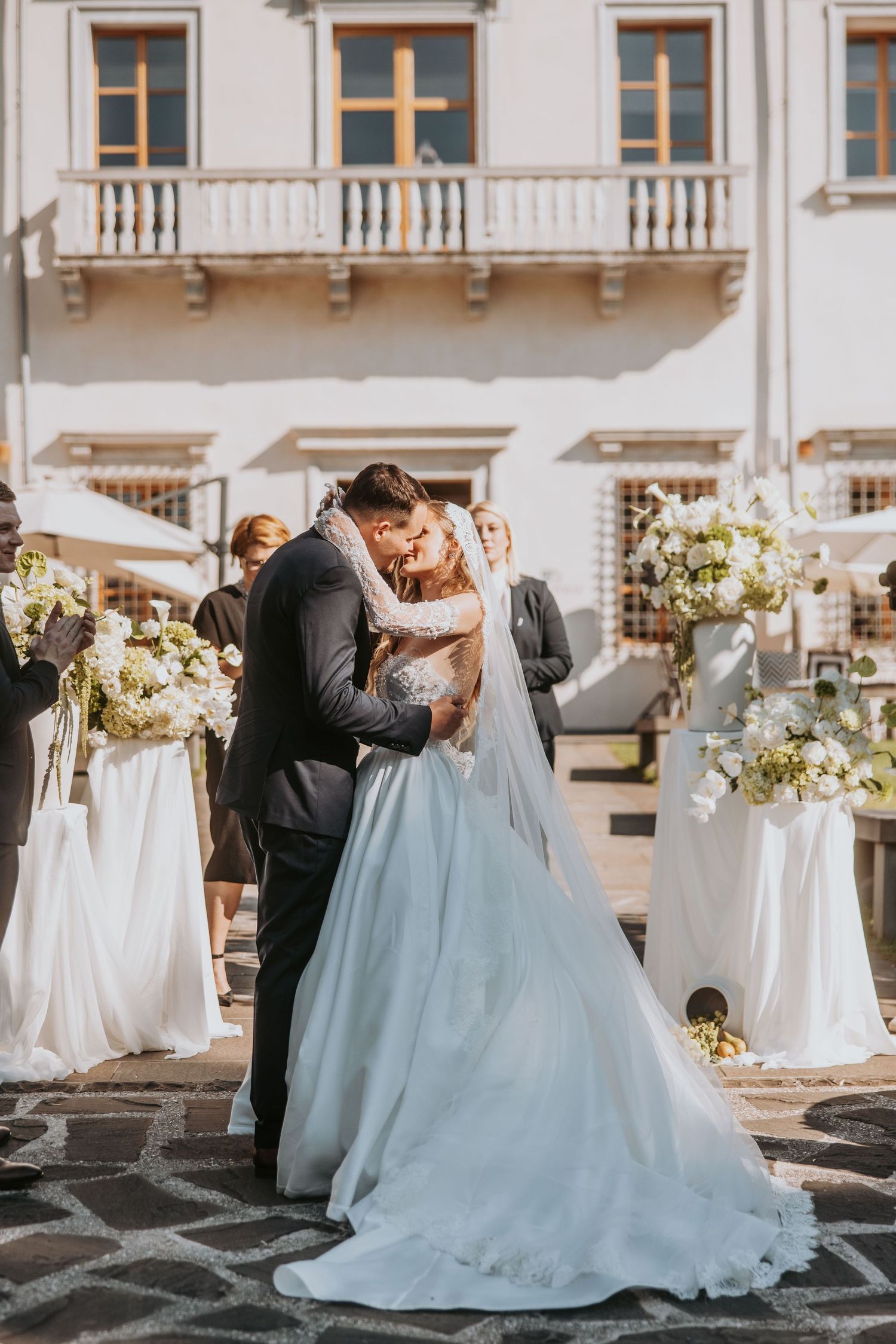 Bride and groom share first kiss in courtyard of elegant European villa during outdoor wedding ceremony.