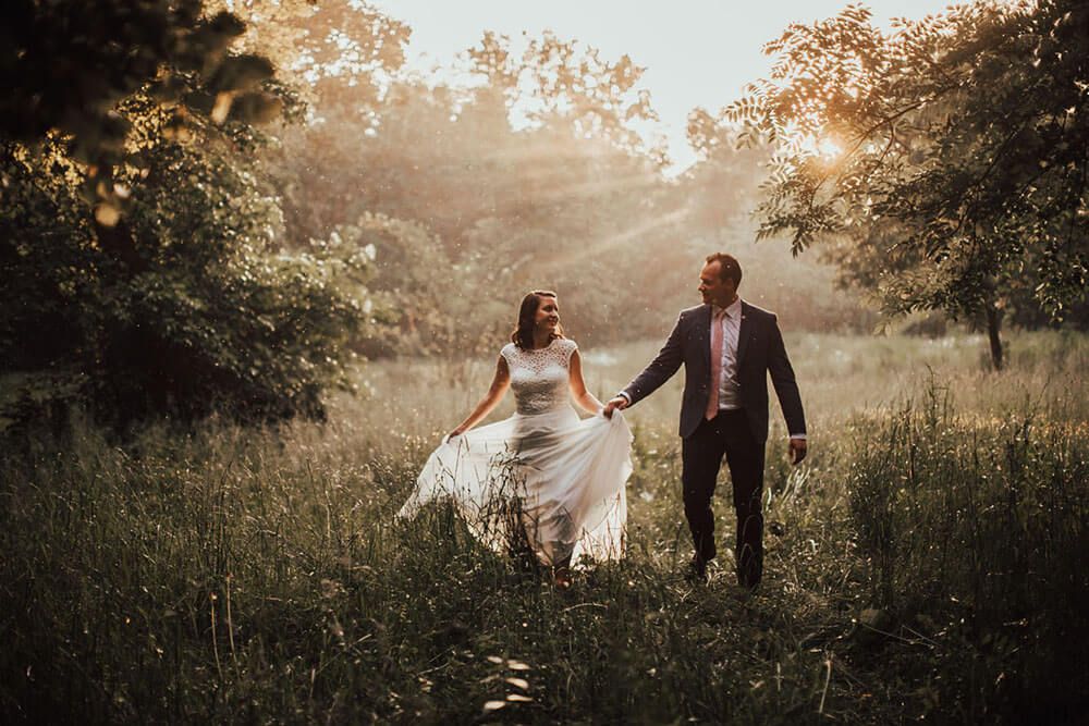 Newlyweds walking hand in hand through a sunlit meadow surrounded by lush green trees at golden hour.