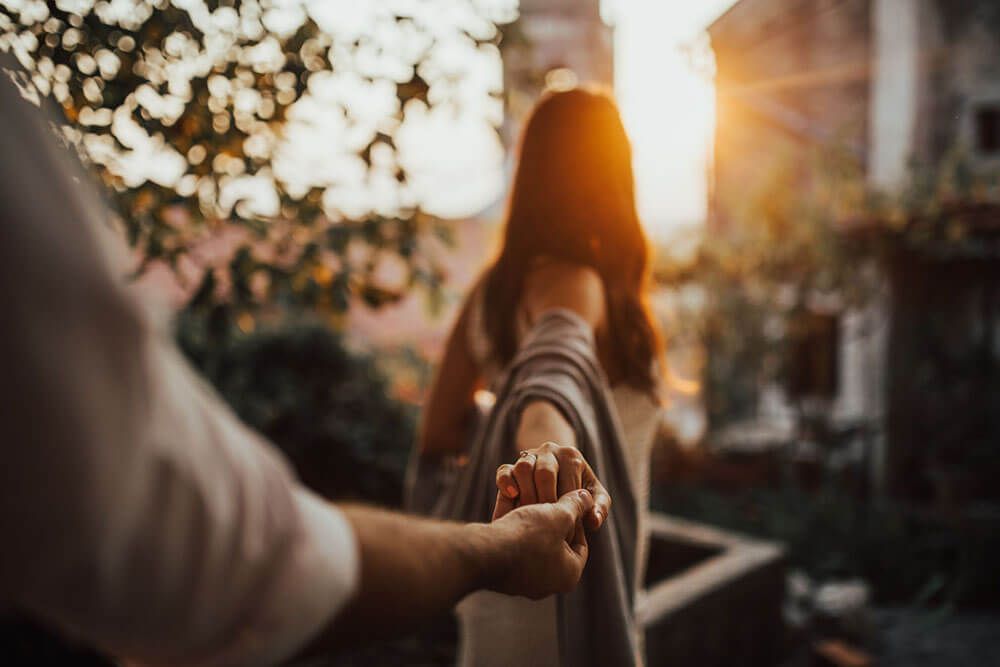 Man holding womans hand leading her through sunlit garden balcony at sunset.