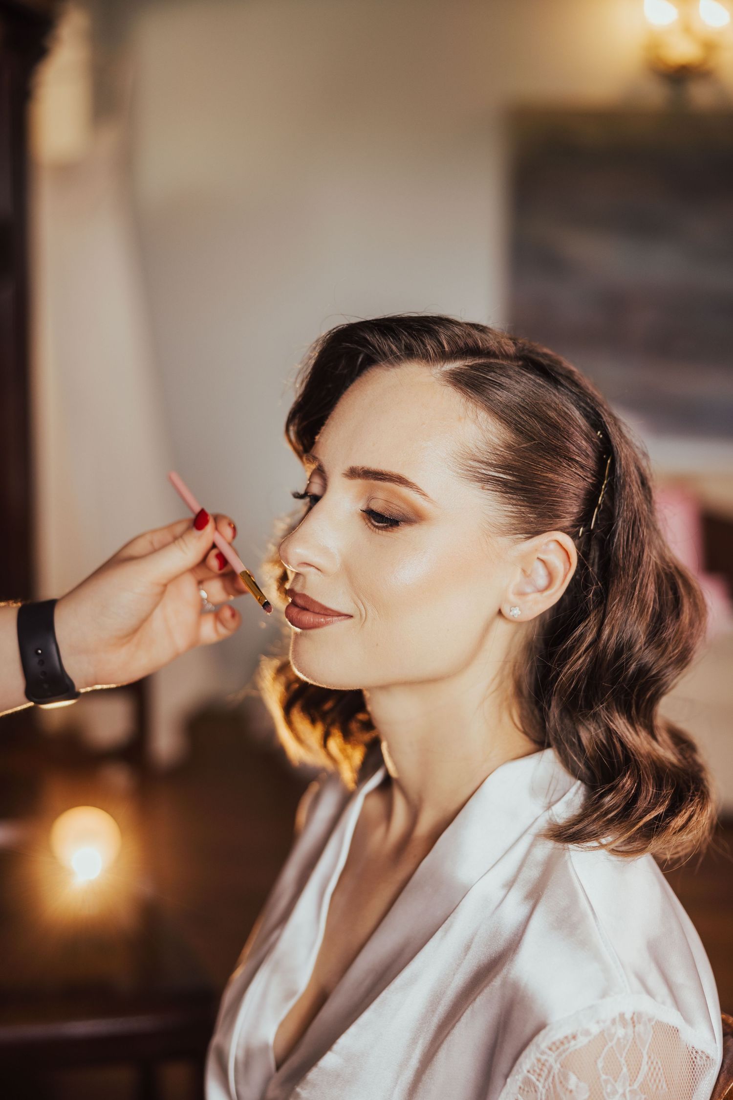 Brunette bride in white robe gets professional makeup applied with brush during wedding preparations.