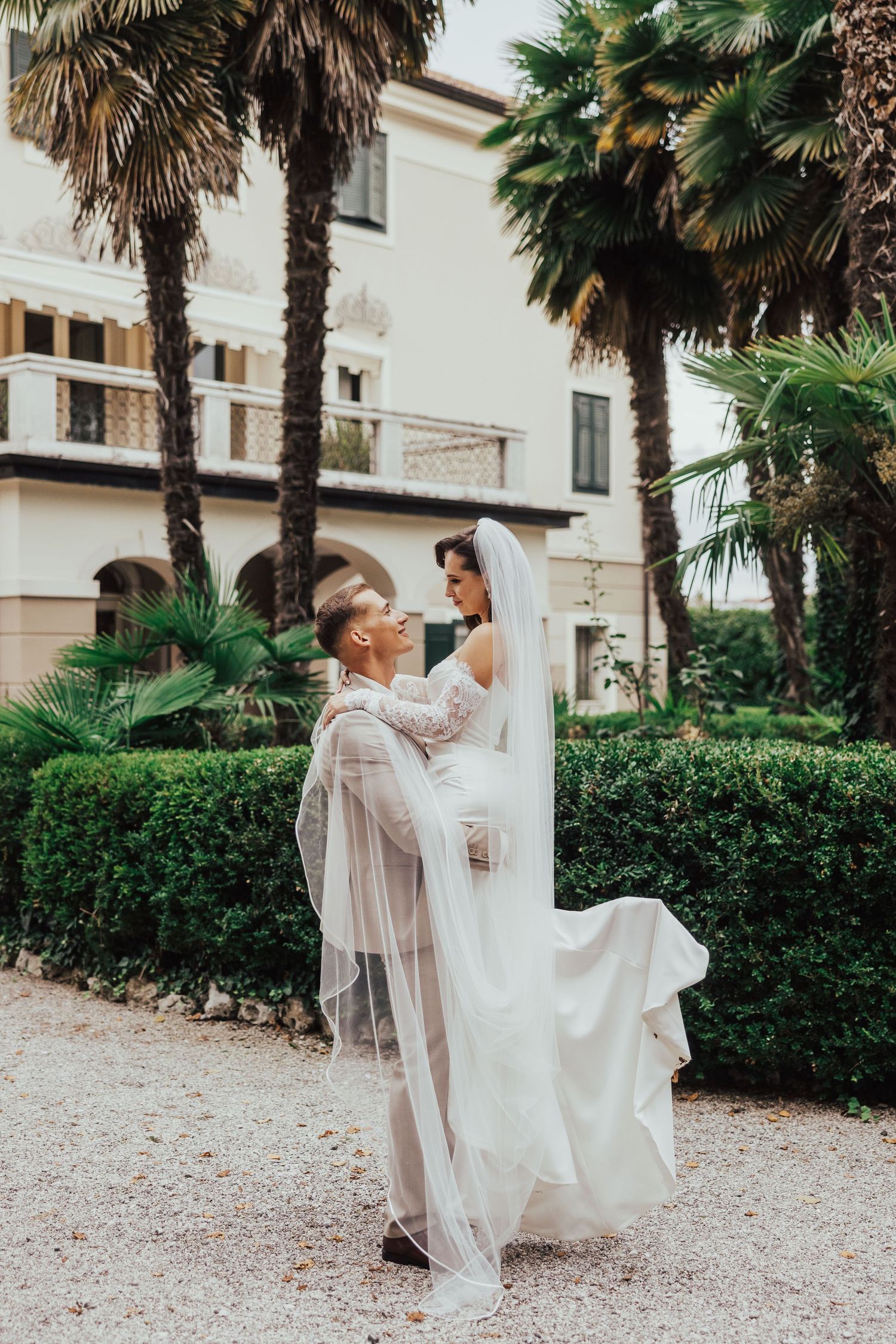 Groom lifting bride in white dress among palm trees at wedding venue.