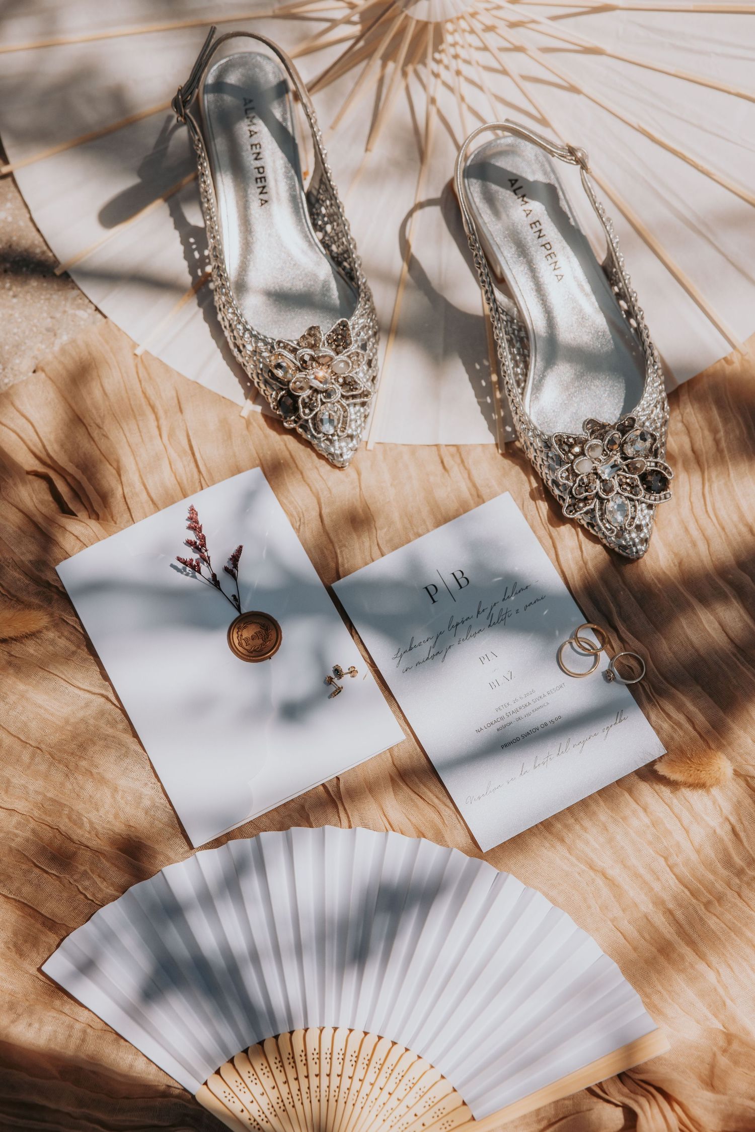 Elegant wedding flat shoes with crystal embellishments beside invitation cards, rings, and a white hand fan on wood.