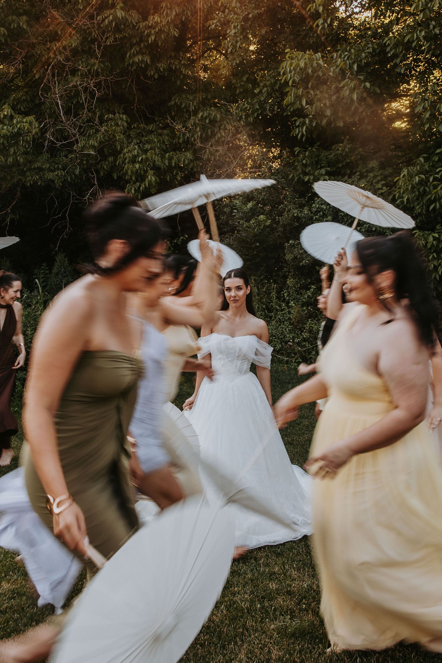 Motion blur photo of bride and bridesmaids dancing outdoors with white parasols at wedding.
