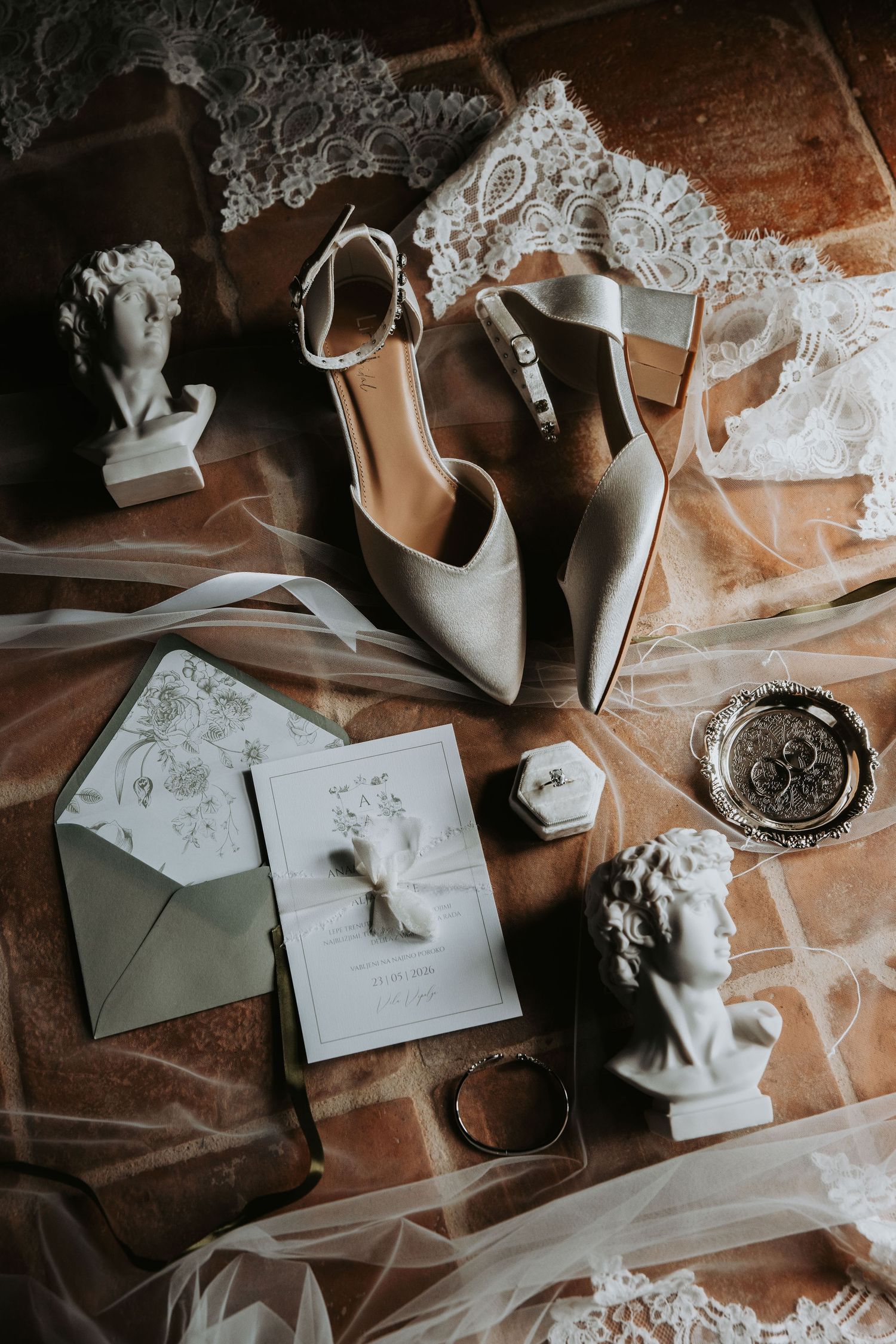 Elegant bridal flat lay featuring ivory heels, wedding invitation, ring box, lace veil, and decorative busts on terracotta tiles.