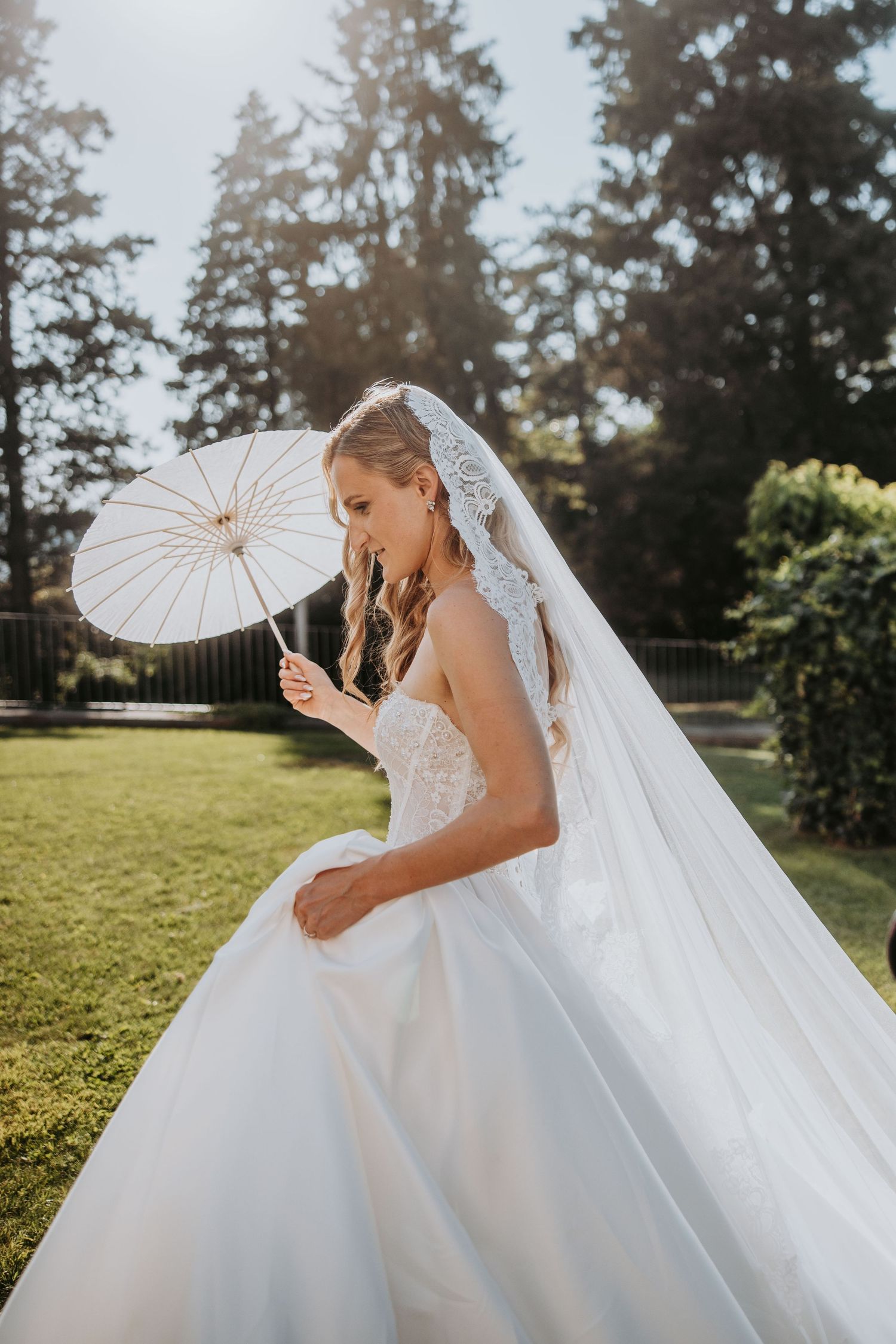 Bride in lace ballgown twirling with white parasol umbrella in sunlit garden on wedding day.