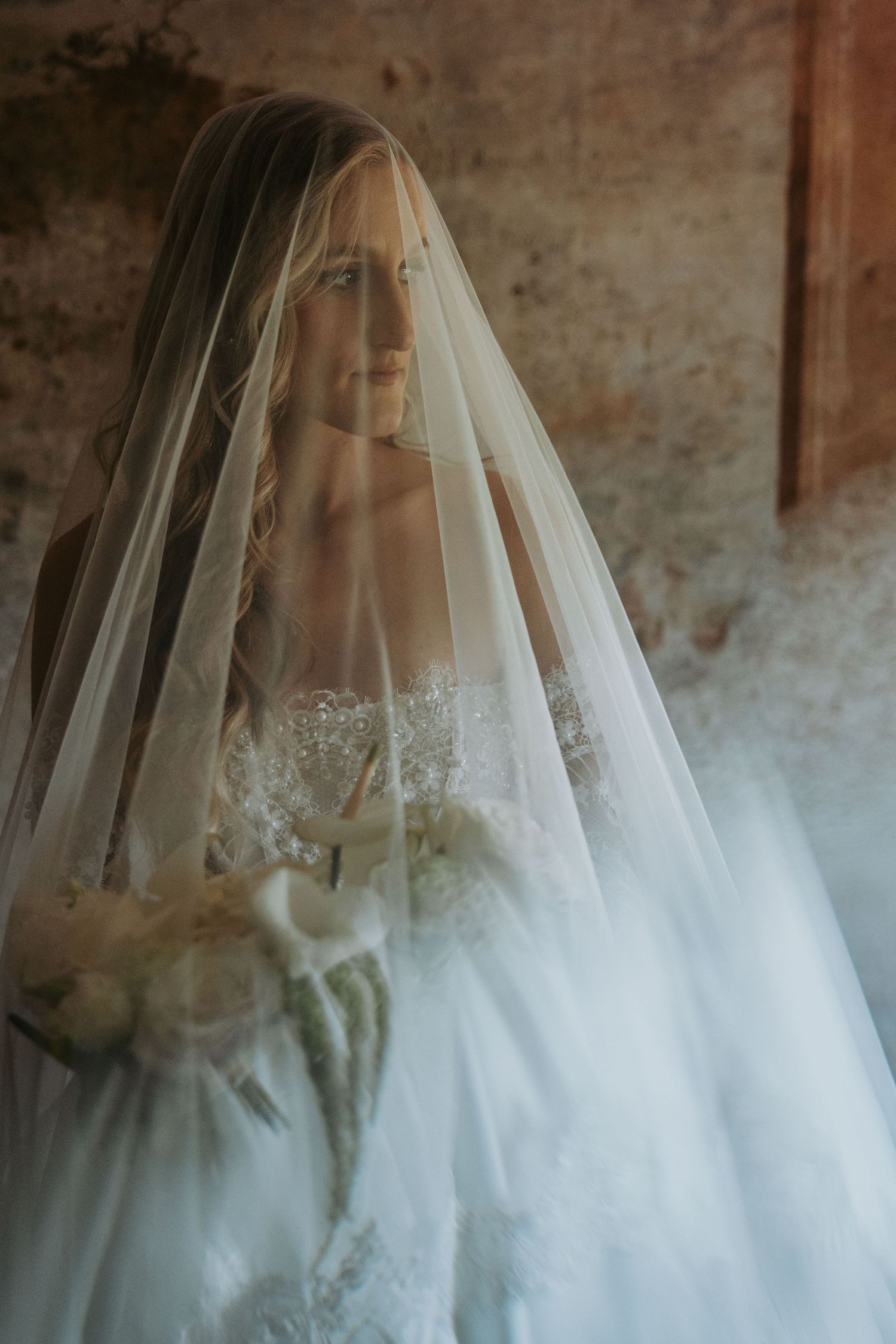Dreamy bridal portrait with blonde bride in white gown holding bouquet beneath flowing veil.
